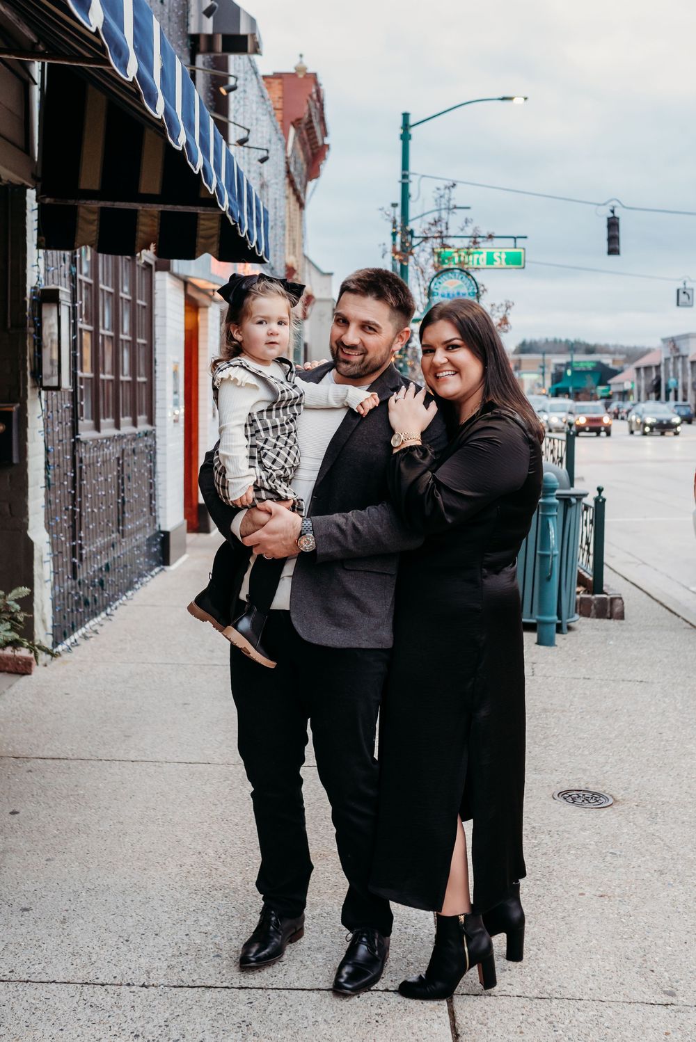 A happy family of three poses on a city sidewalk, man holding toddler, woman in black dress beside them.