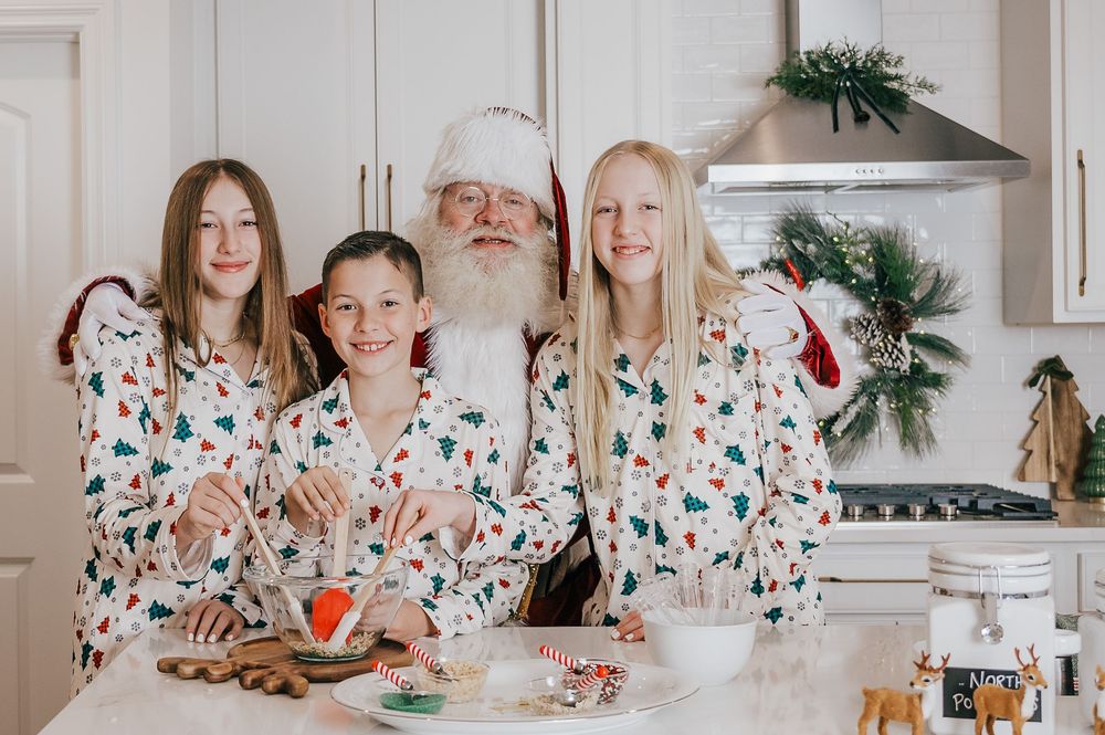 Santa Claus poses with three children in matching Christmas pajamas while baking holiday cookies in a festive kitchen.