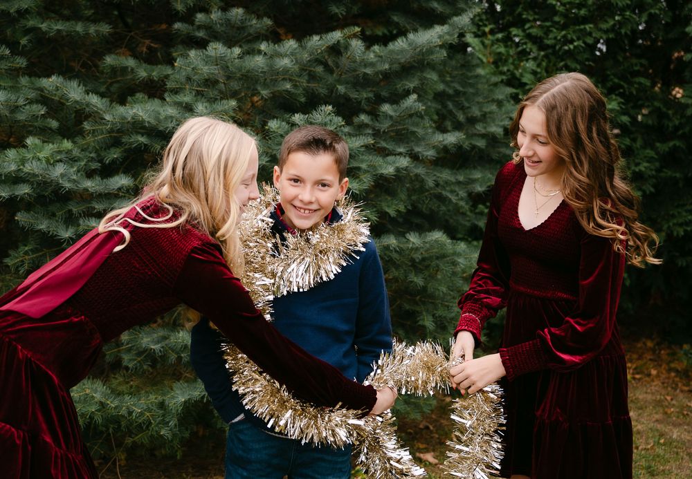 Two girls in red velvet dresses decorating a boy with gold tinsel garland outdoors near evergreen trees.