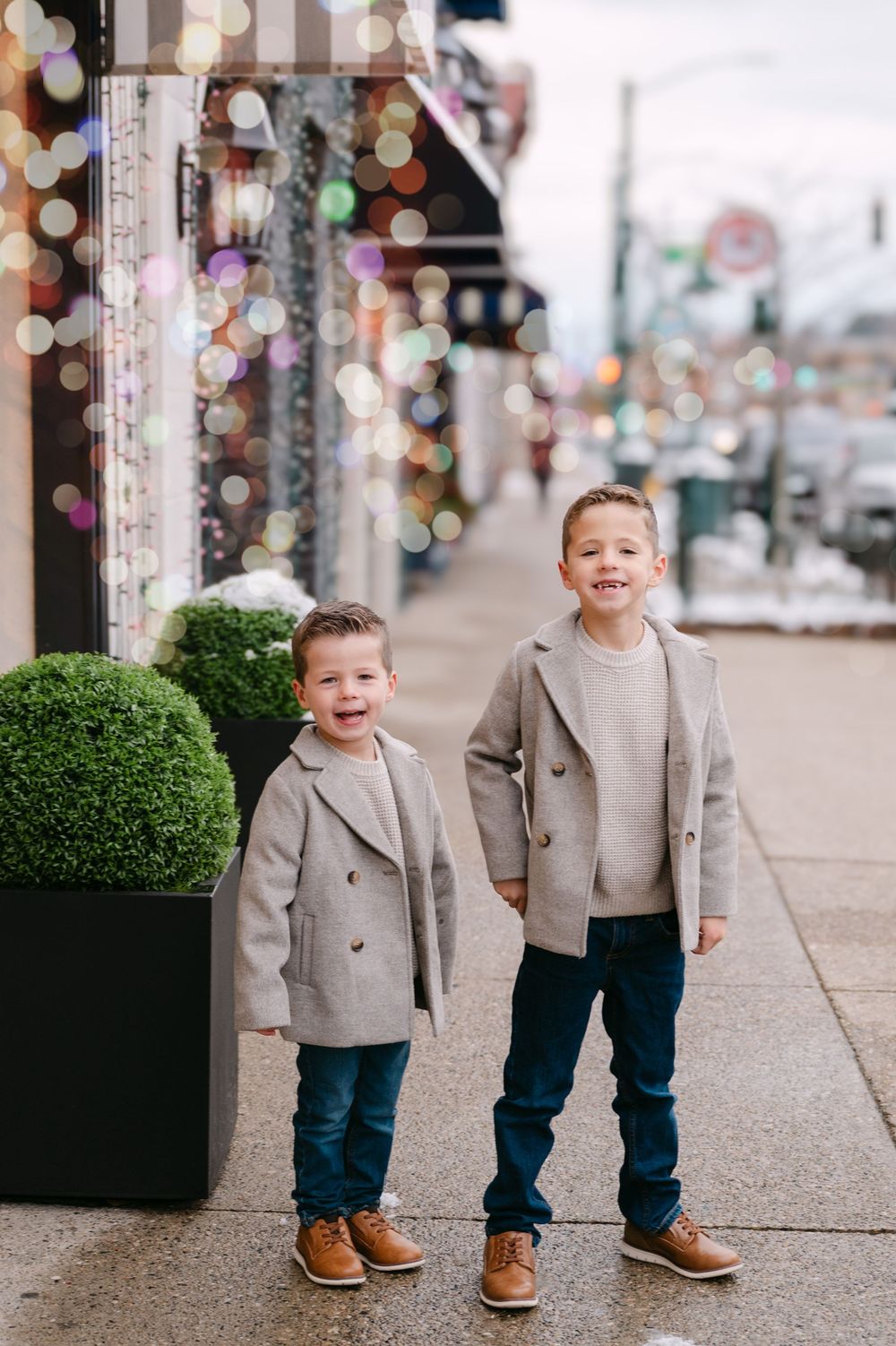 Two young boys in matching gray coats and jeans smile on a festive sidewalk with bokeh holiday lights.