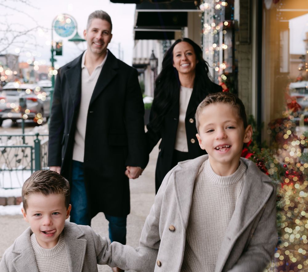 A happy family of four poses on a festive small-town sidewalk decorated with holiday lights during winter.