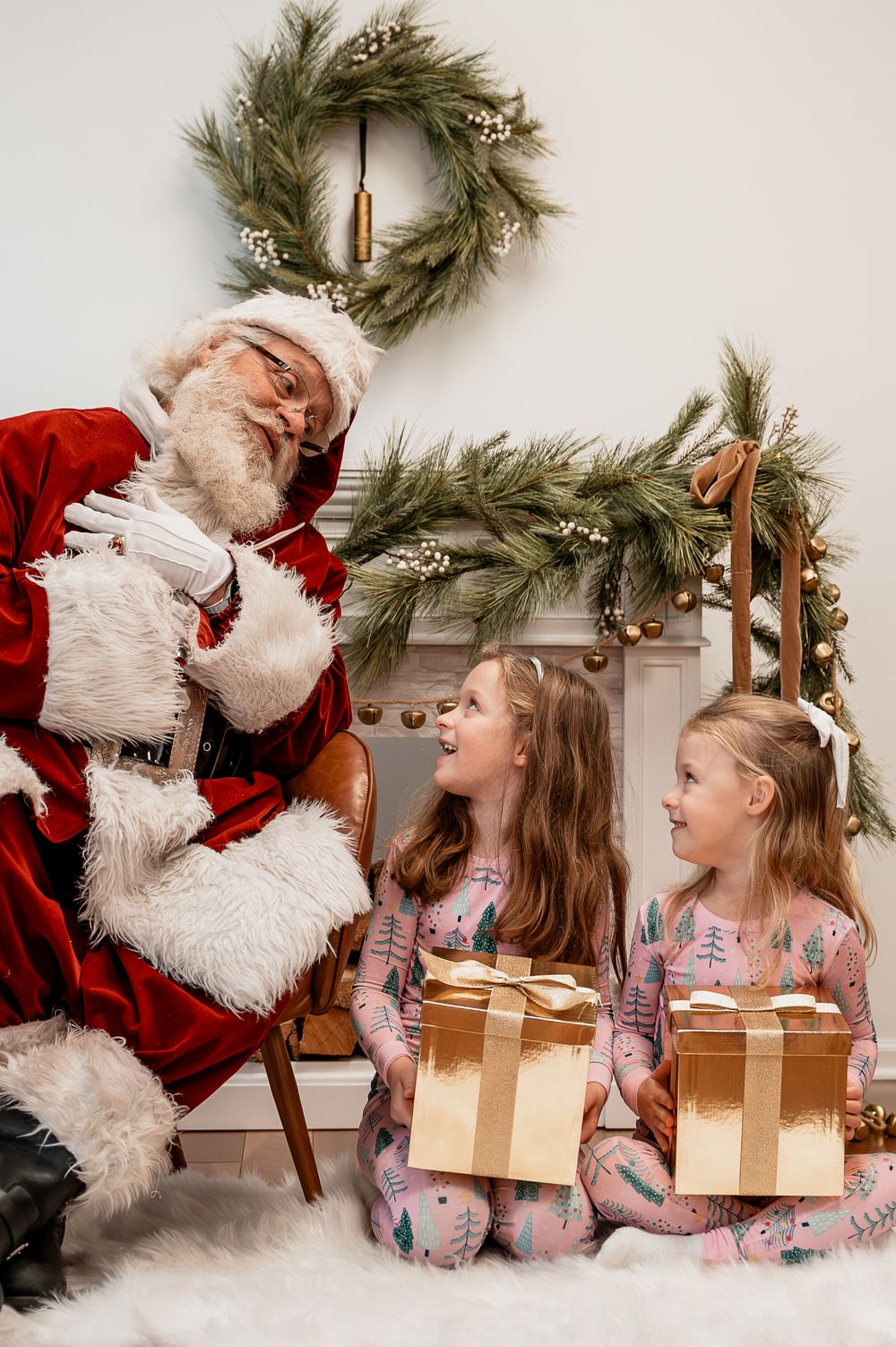 Two girls in Christmas pajamas hold gold gifts while sitting with Santa Claus near a festive wreath and garland.