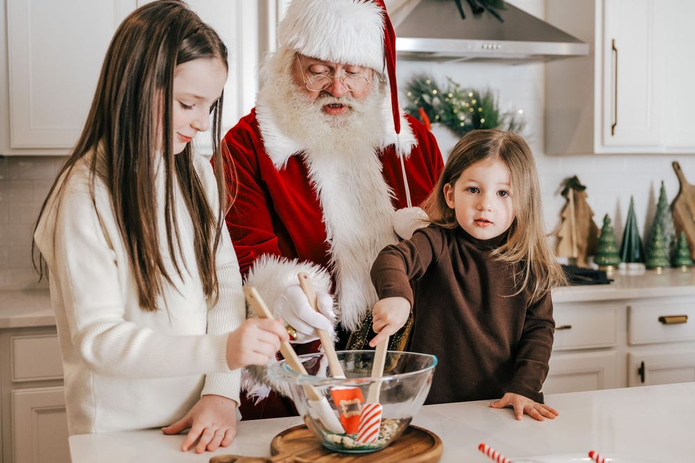 Santa Claus baking cookies with two young girls in a festive white kitchen during Christmas.