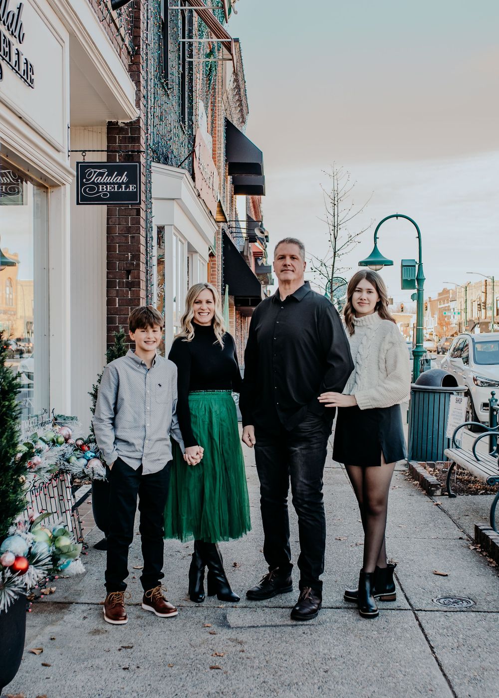 Family of four poses on a charming downtown sidewalk near festive holiday decorations outside a boutique shop.