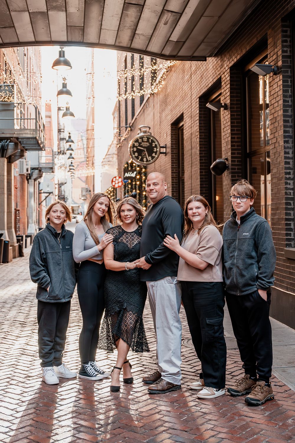 Family of six posing together in a charming brick alley decorated with string lights and a vintage clock.