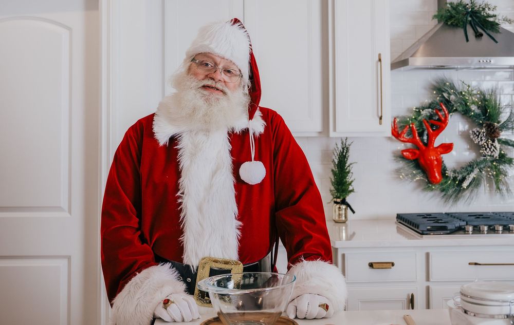 Santa Claus standing in a decorated kitchen holding a gift box, surrounded by Christmas decorations.