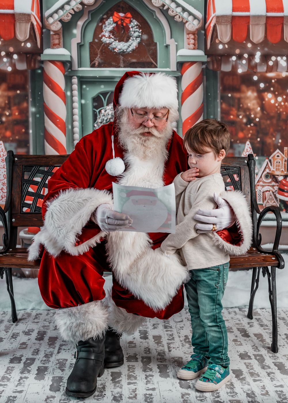 Santa Claus reads a letter with a young boy standing beside him in a festive Christmas photo studio setting.