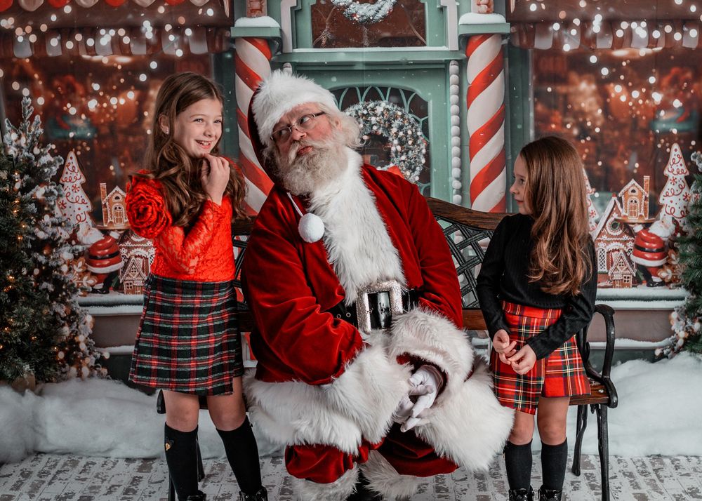 Two girls in plaid skirts pose with Santa Claus in a festive Christmas workshop setting with gingerbread decorations.