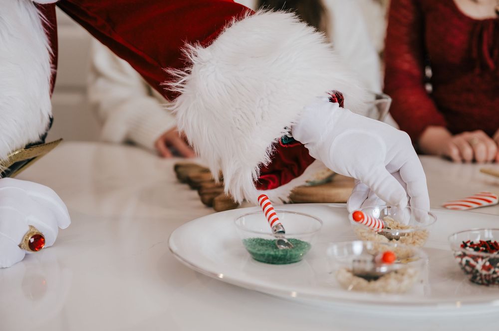 Santa Claus decorating Christmas cookies with candy cane and green sprinkles on a white plate.