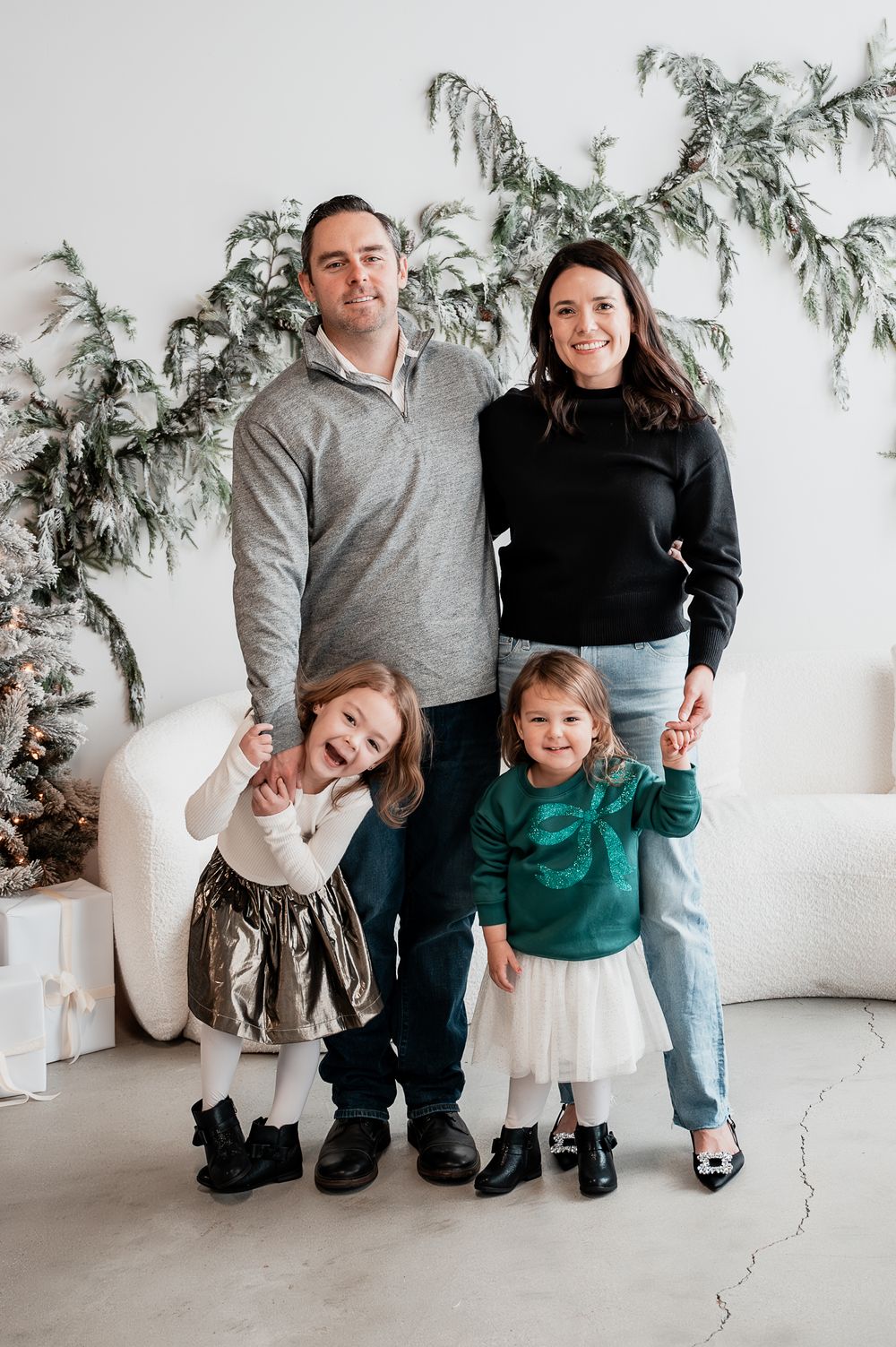 Family of four posing for a festive Christmas photo with holiday greenery and gifts in a bright white studio.
