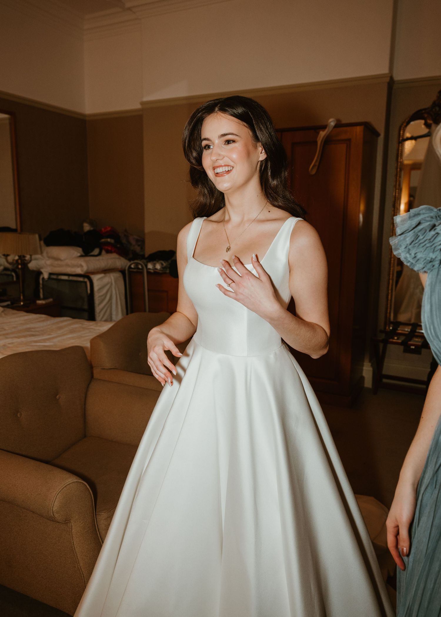 Smiling bride in elegant white satin ball gown dress during wedding preparation in a warmly lit room.