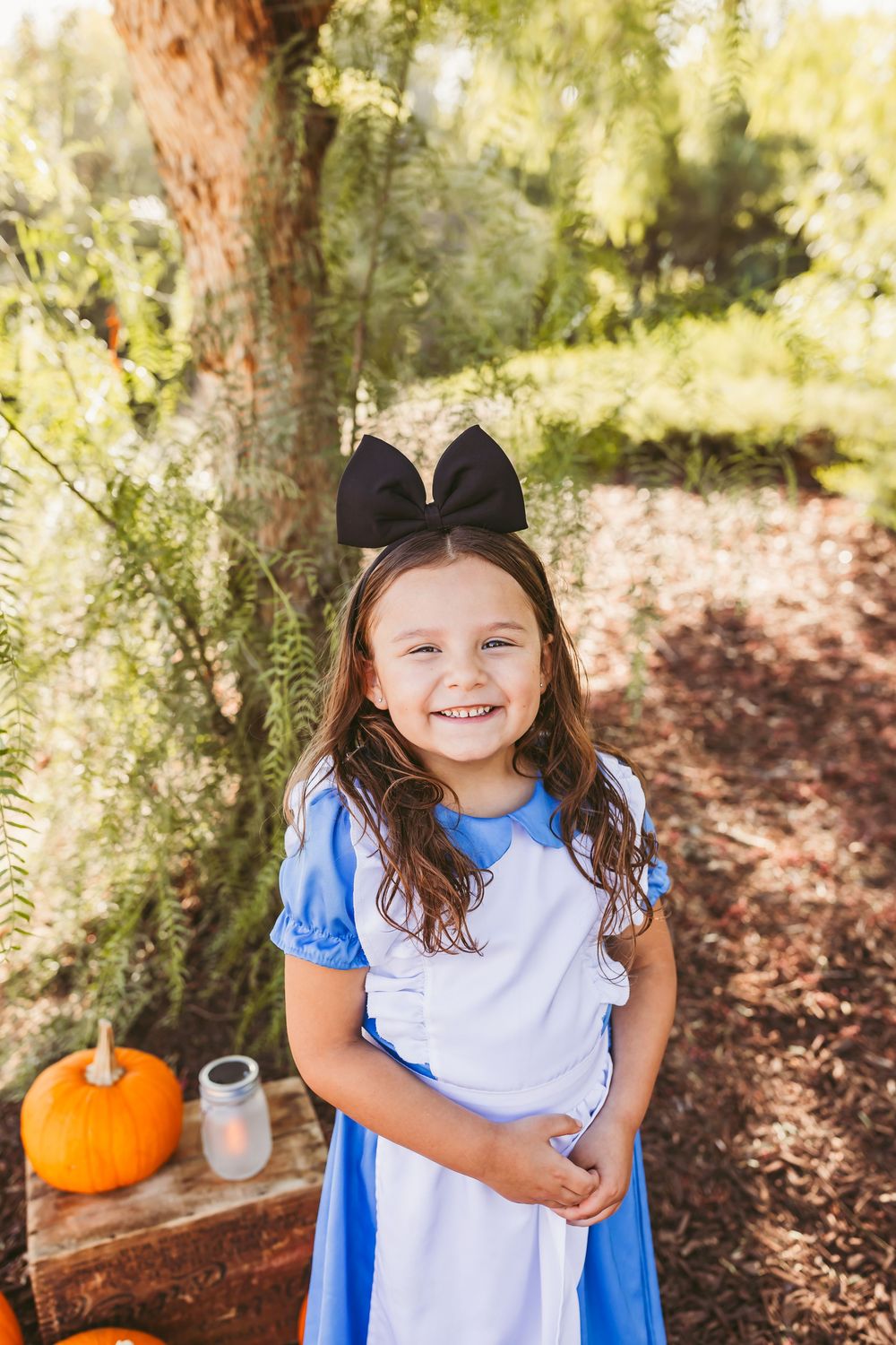 Young girl in Alice in Wonderland costume with black bow, smiling outdoors near pumpkins and greenery.