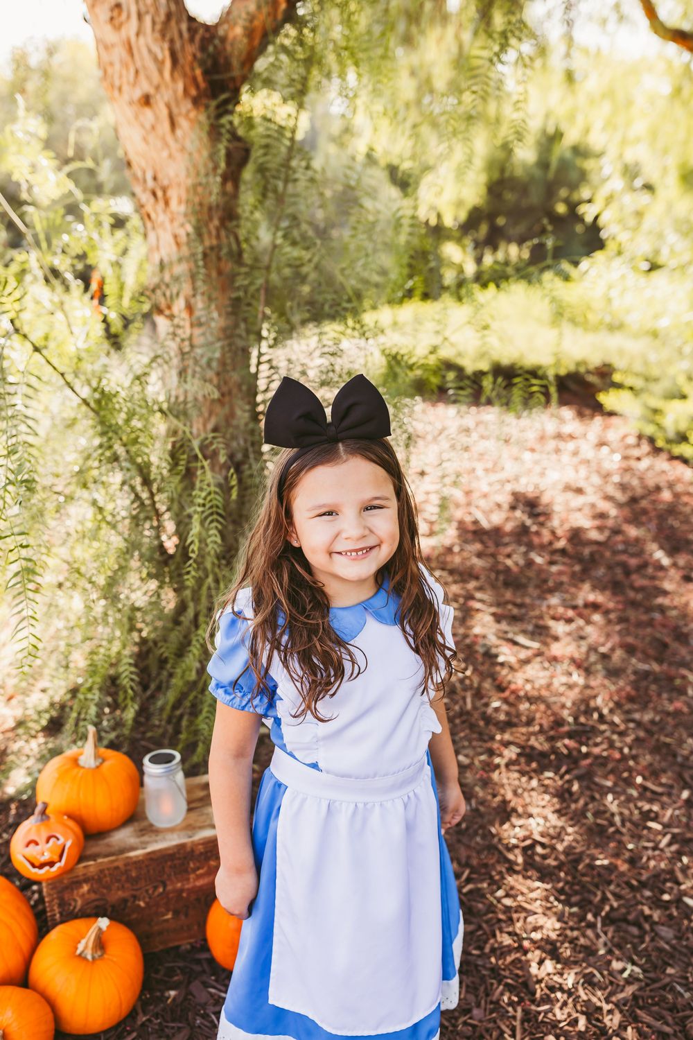 Young girl in Alice in Wonderland costume with black bow smiles near Halloween pumpkins outdoors.