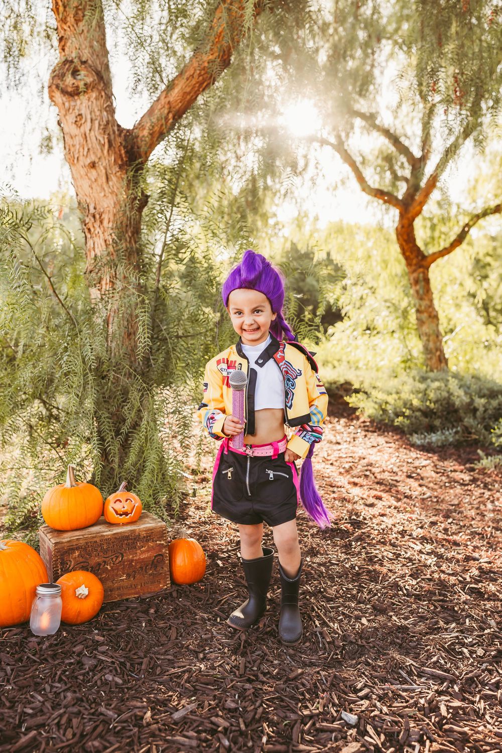 Girl in purple wig and colorful Halloween costume holds prop near pumpkins outdoors in sunny forest setting.