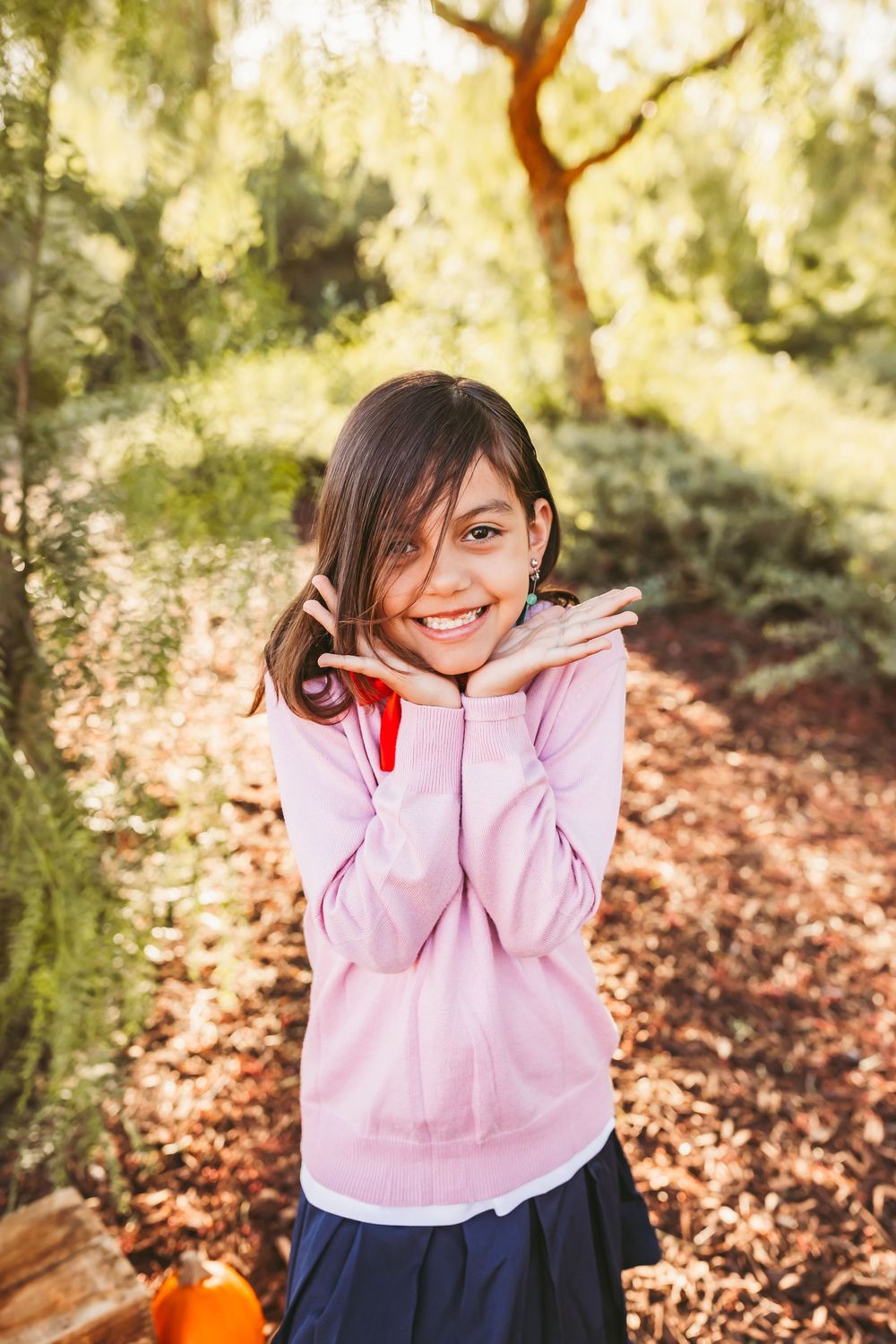Smiling young girl in pink hoodie posing with hands on cheeks in a sunlit autumn forest setting.