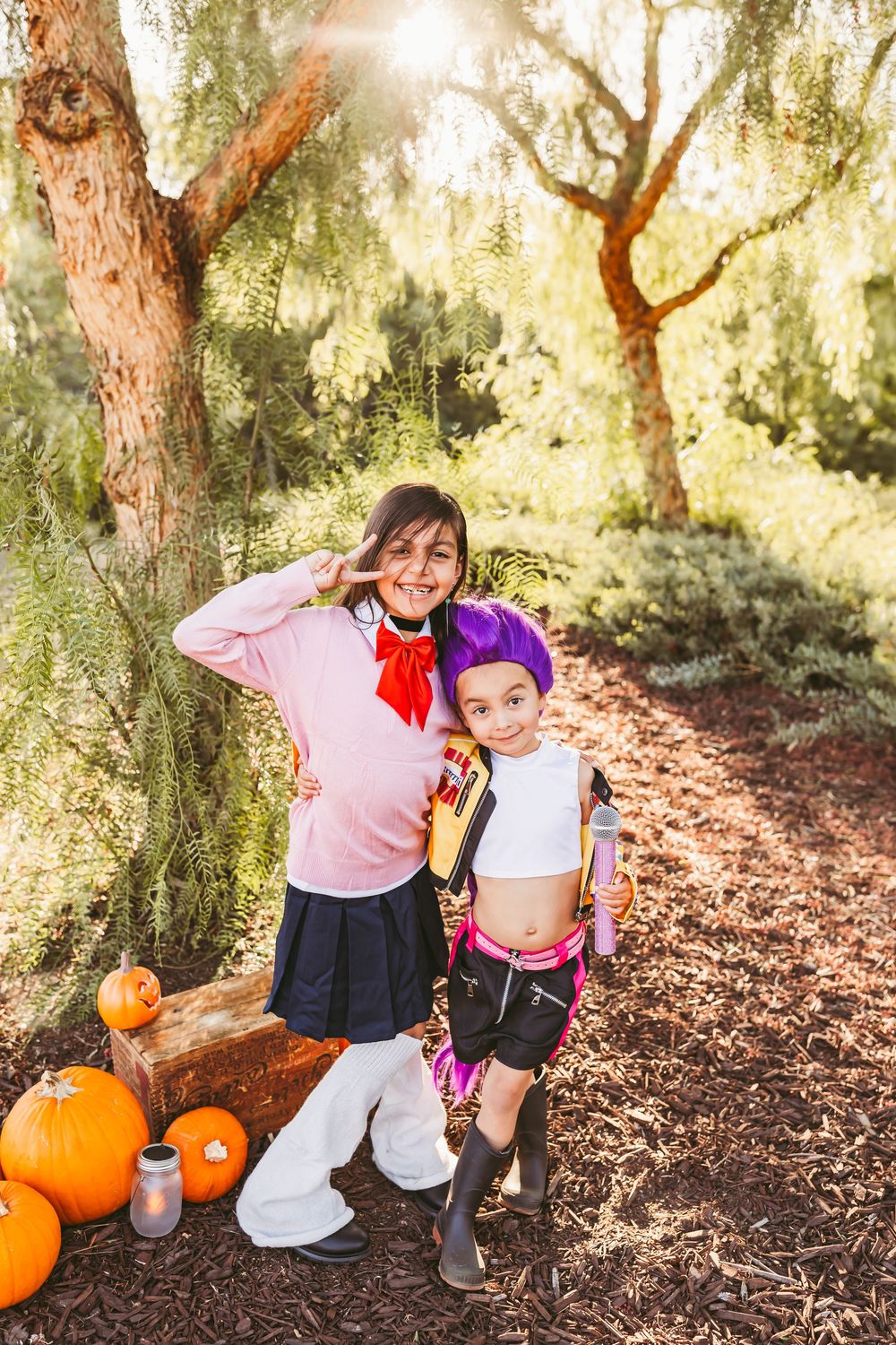 Two children in Halloween costumes pose outdoors among pumpkins and trees in warm sunlight.
