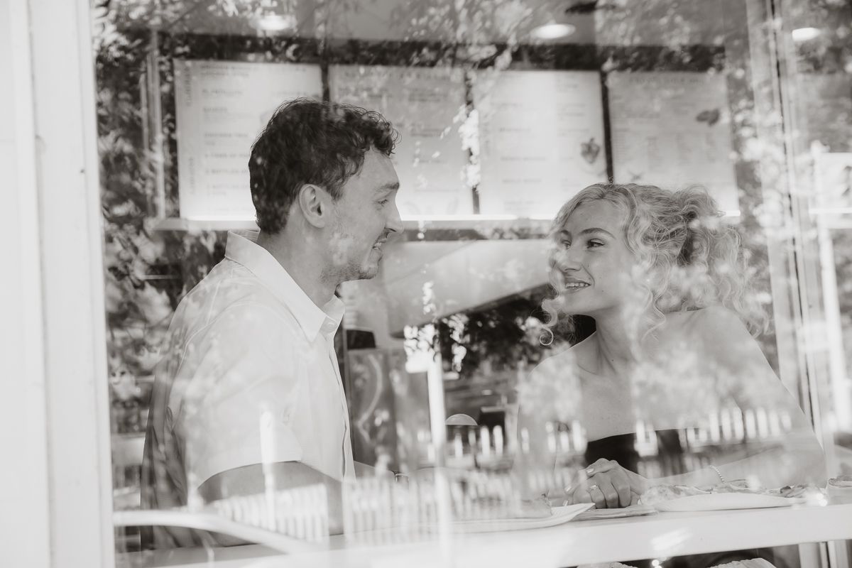 Black and white photo of a smiling couple sitting together at a cafe table, viewed through a window.