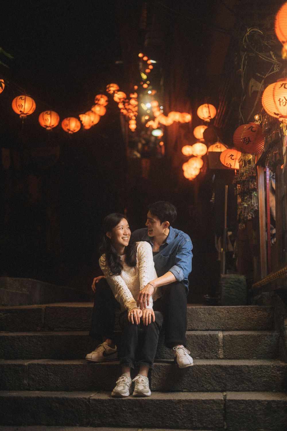 Couple sitting on steps under glowing red lanterns at night in a romantic Asian street setting.