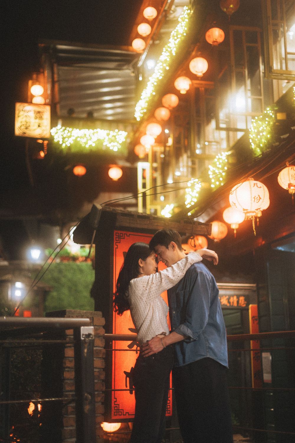Couple embracing romantically at night under glowing red lanterns in a traditional Asian street setting.