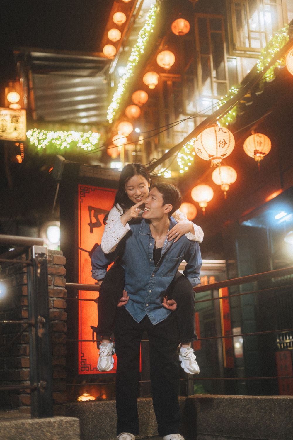 Couple sharing a joyful piggyback ride at night under glowing red lanterns in a festive Asian street setting.