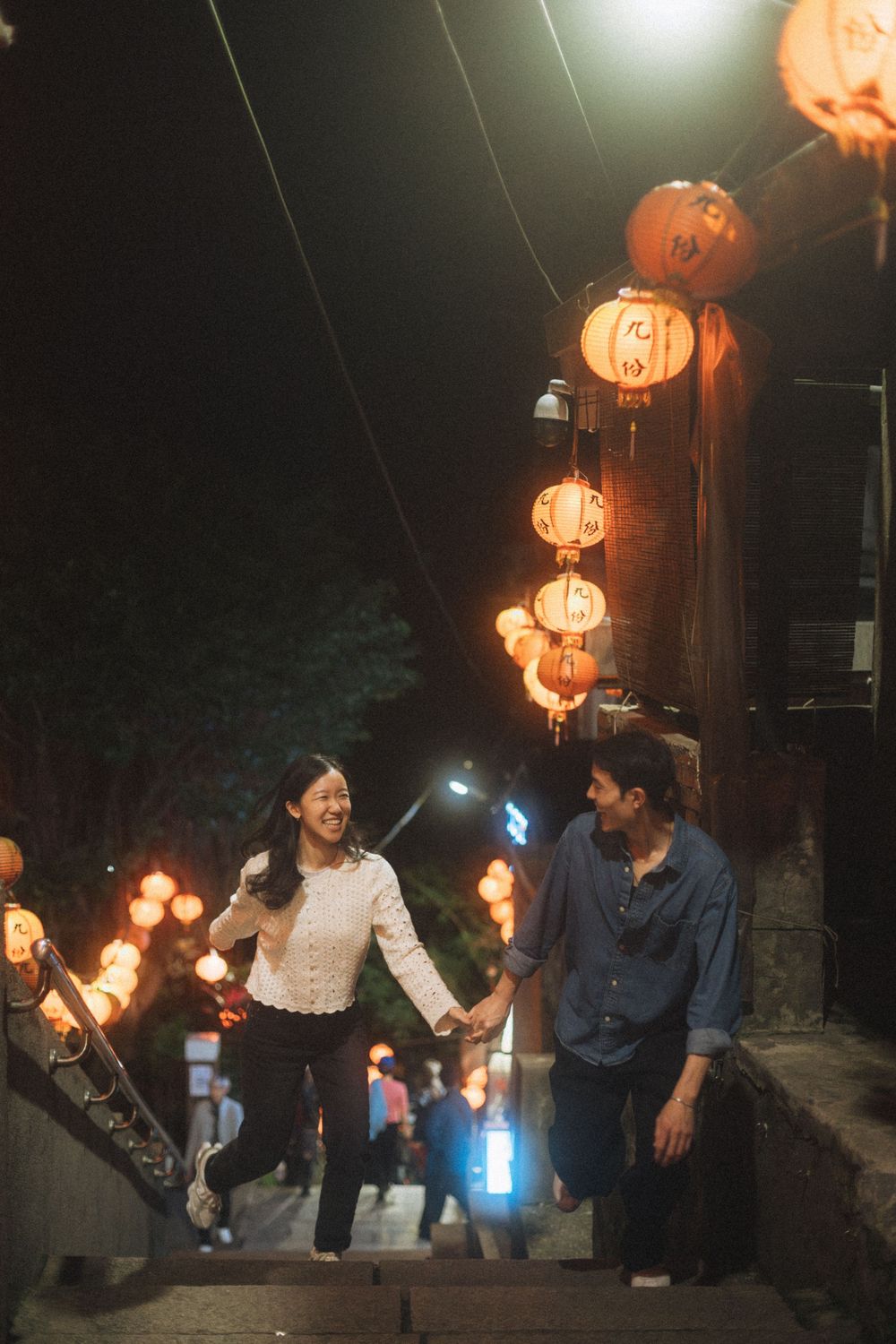 Couple holding hands on lantern-lit steps at night in an Asian night market setting.