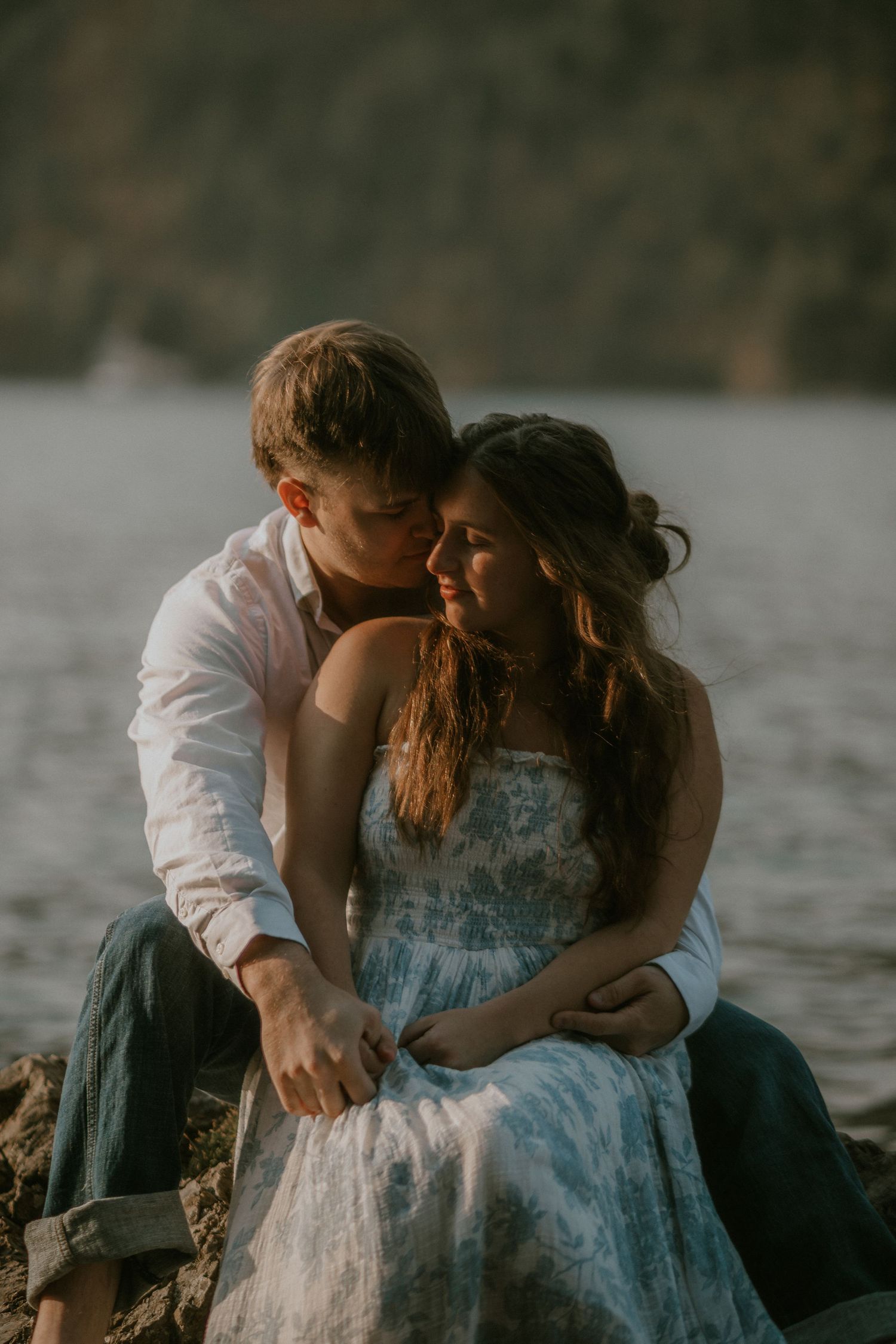 A couple shares a tender moment sitting by a lake, the man kissing the womans cheek as she smiles in golden light.