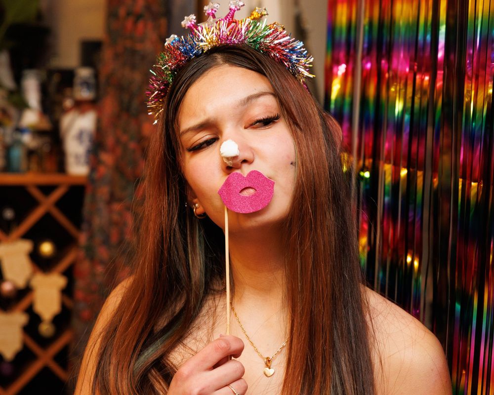 Young woman wearing colorful tinsel crown holds pink lips prop at festive party photo booth.