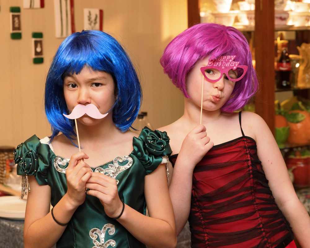 Two girls in colorful wigs holding birthday party photo booth props, wearing festive dresses at a birthday celebration.