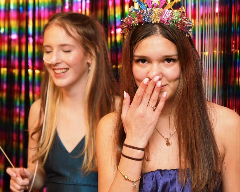 Two teenage girls celebrating at a colorful party, one wearing a floral crown, posing in front of a rainbow tinsel backdrop.