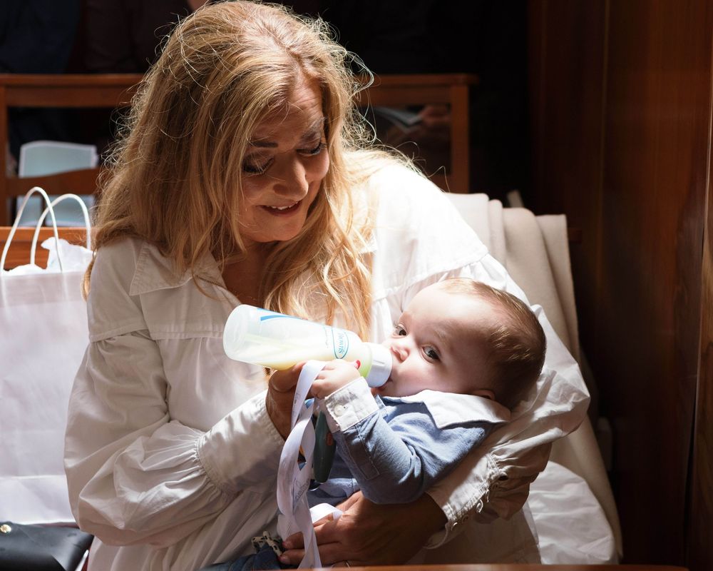 Blonde woman in white shirt bottle feeding a baby dressed in blue outfit indoors.