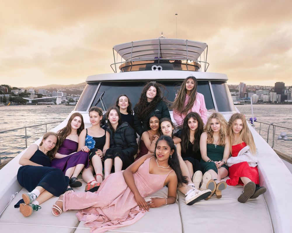 Group of young women posing on a luxury yacht deck with city skyline and golden sky backdrop.
