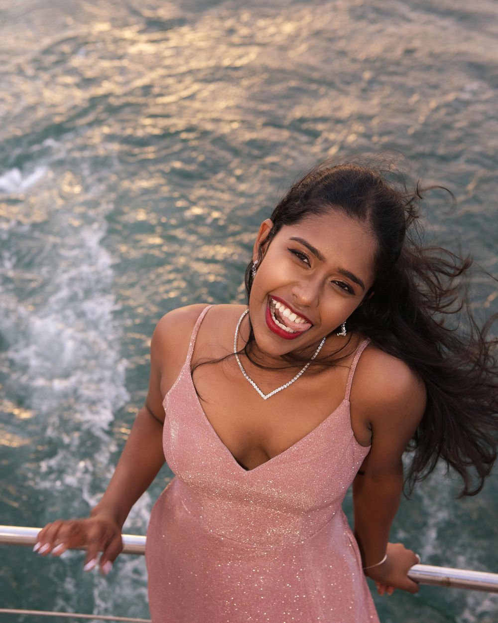 A smiling young woman in a pink sparkly dress leans on a railing with ocean waves in the background.