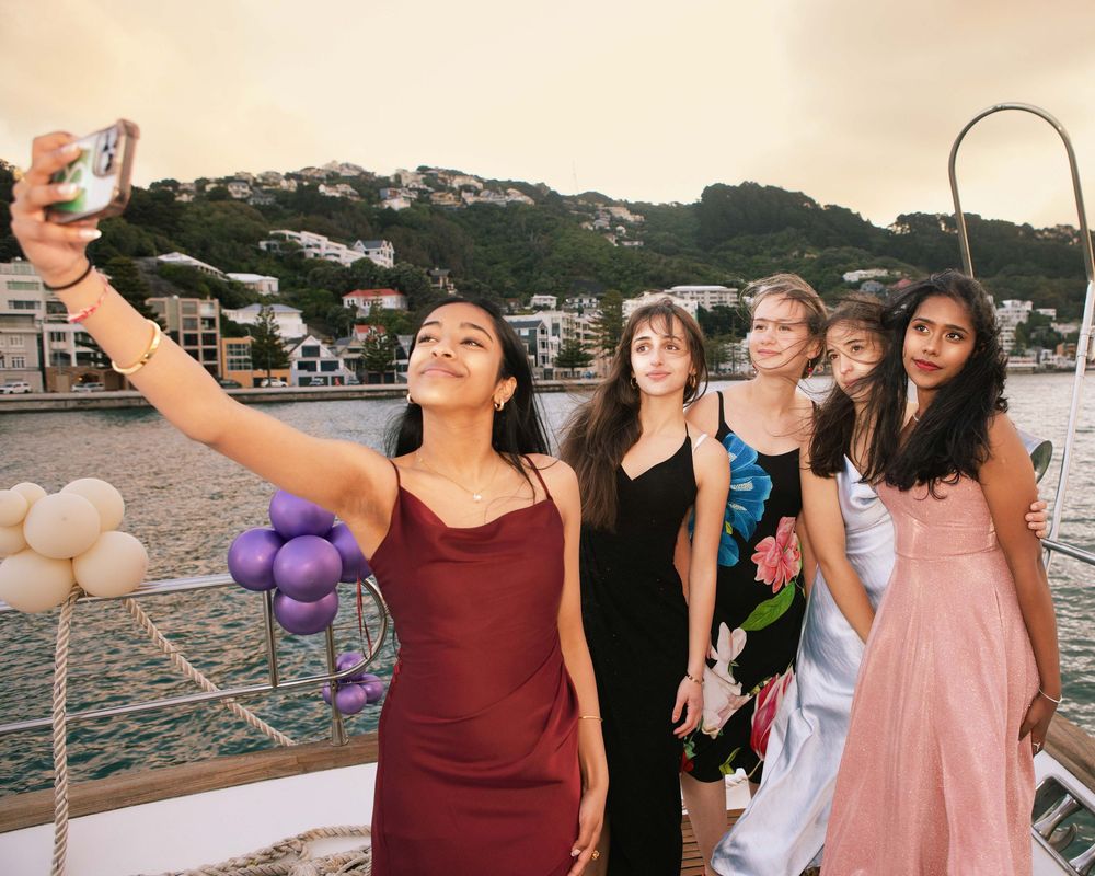 Group of teenage girls in formal dresses taking a selfie on a boat deck with harbor views.