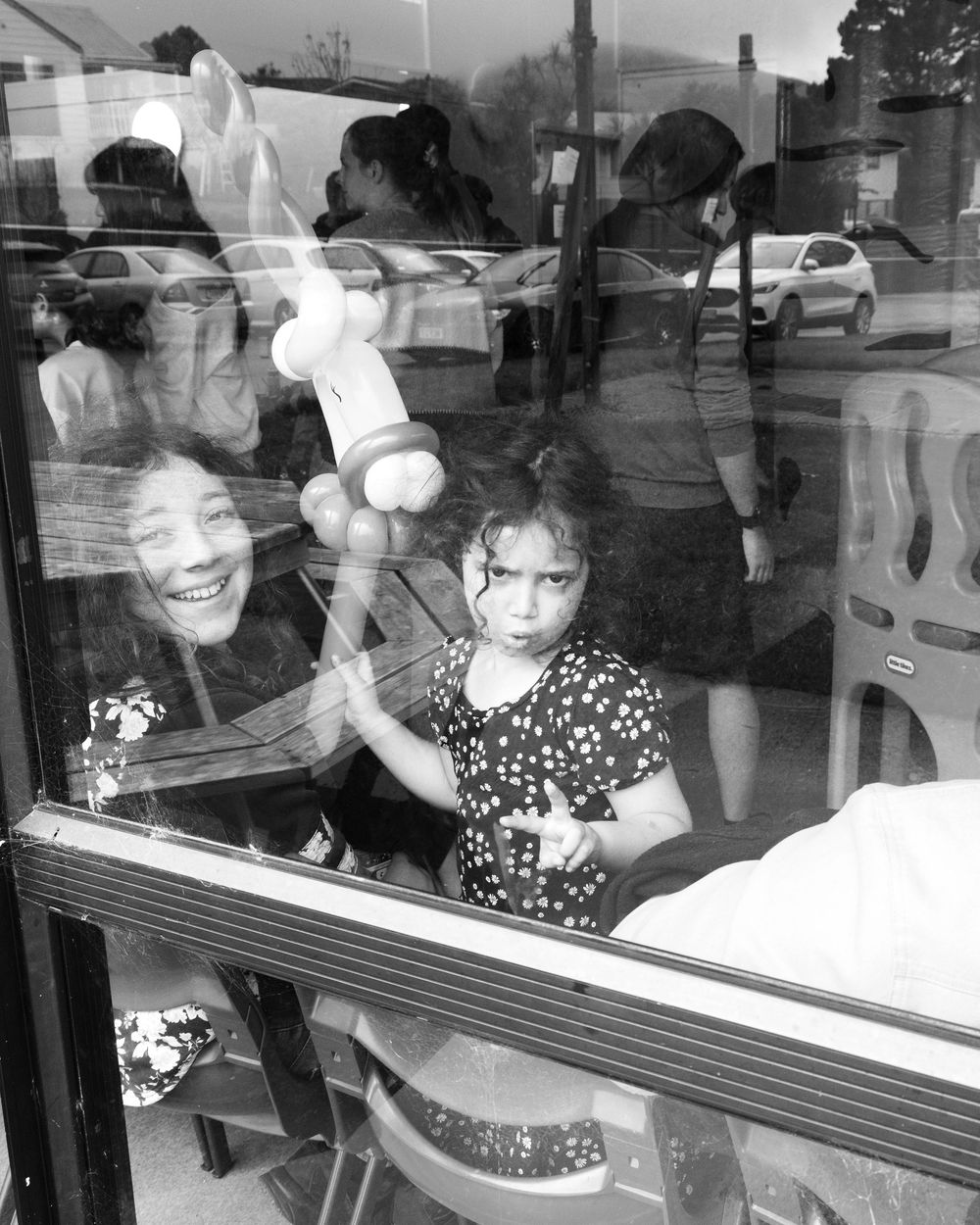 Black and white photo of a young girl and smiling woman seen through a window, with reflections of a street scene.