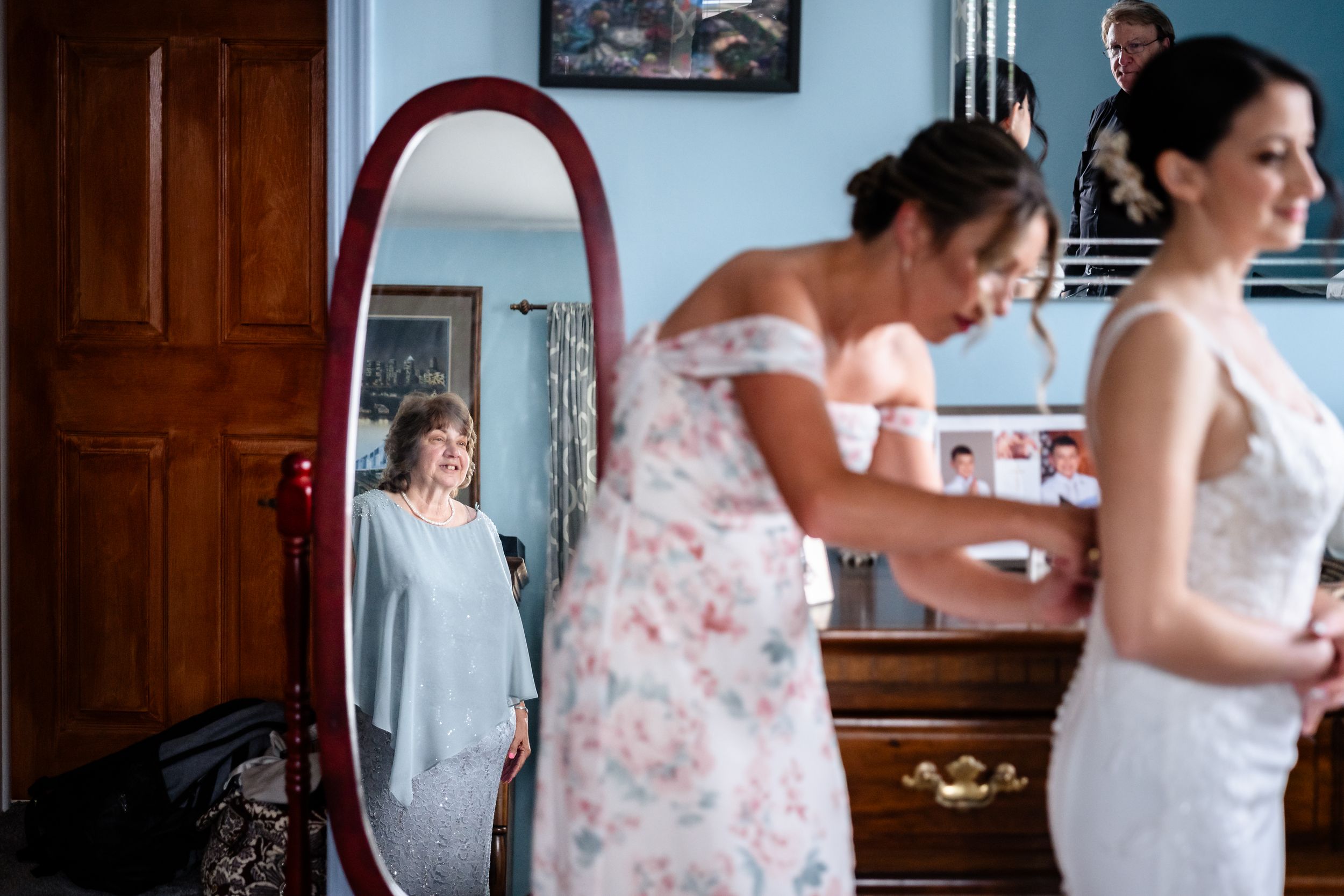 Mother watches daughter in wedding dress being helped with gown in mirror reflection.