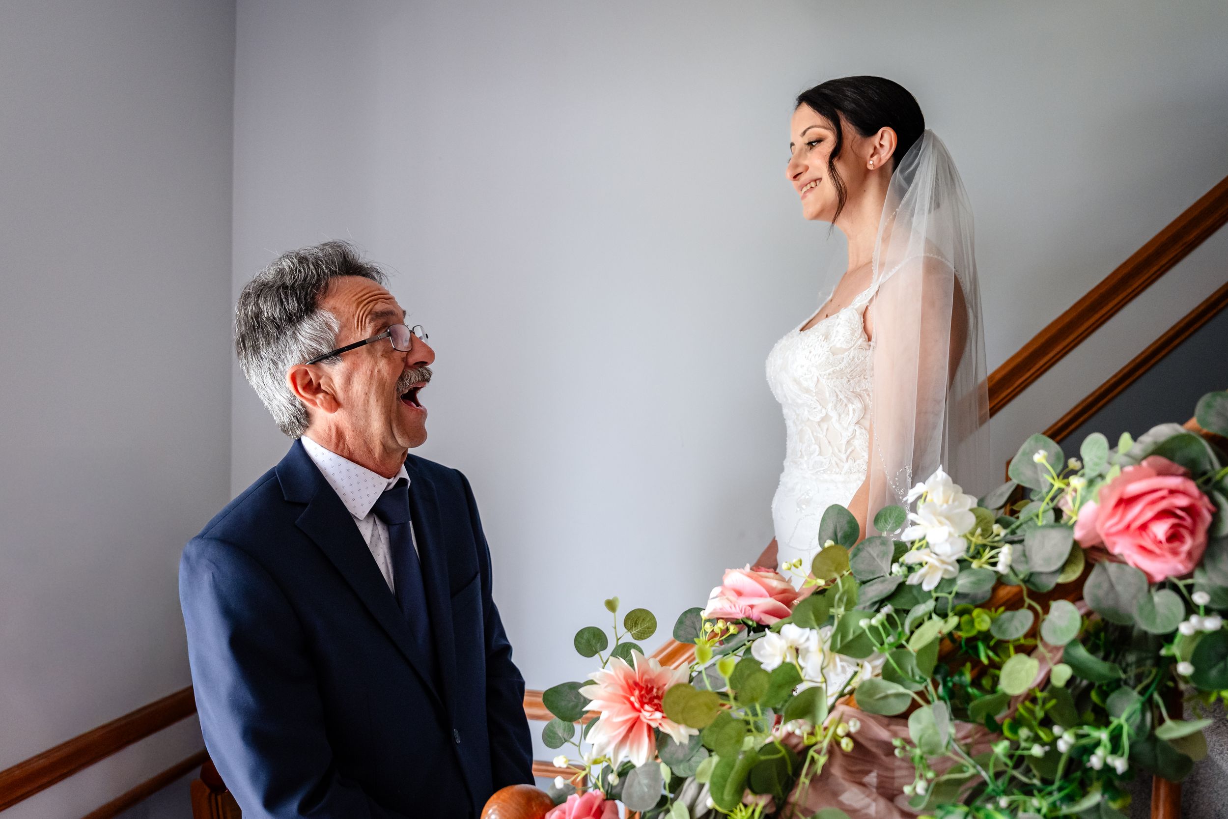 Emotional father smiles up at daughter in wedding dress and veil on staircase.