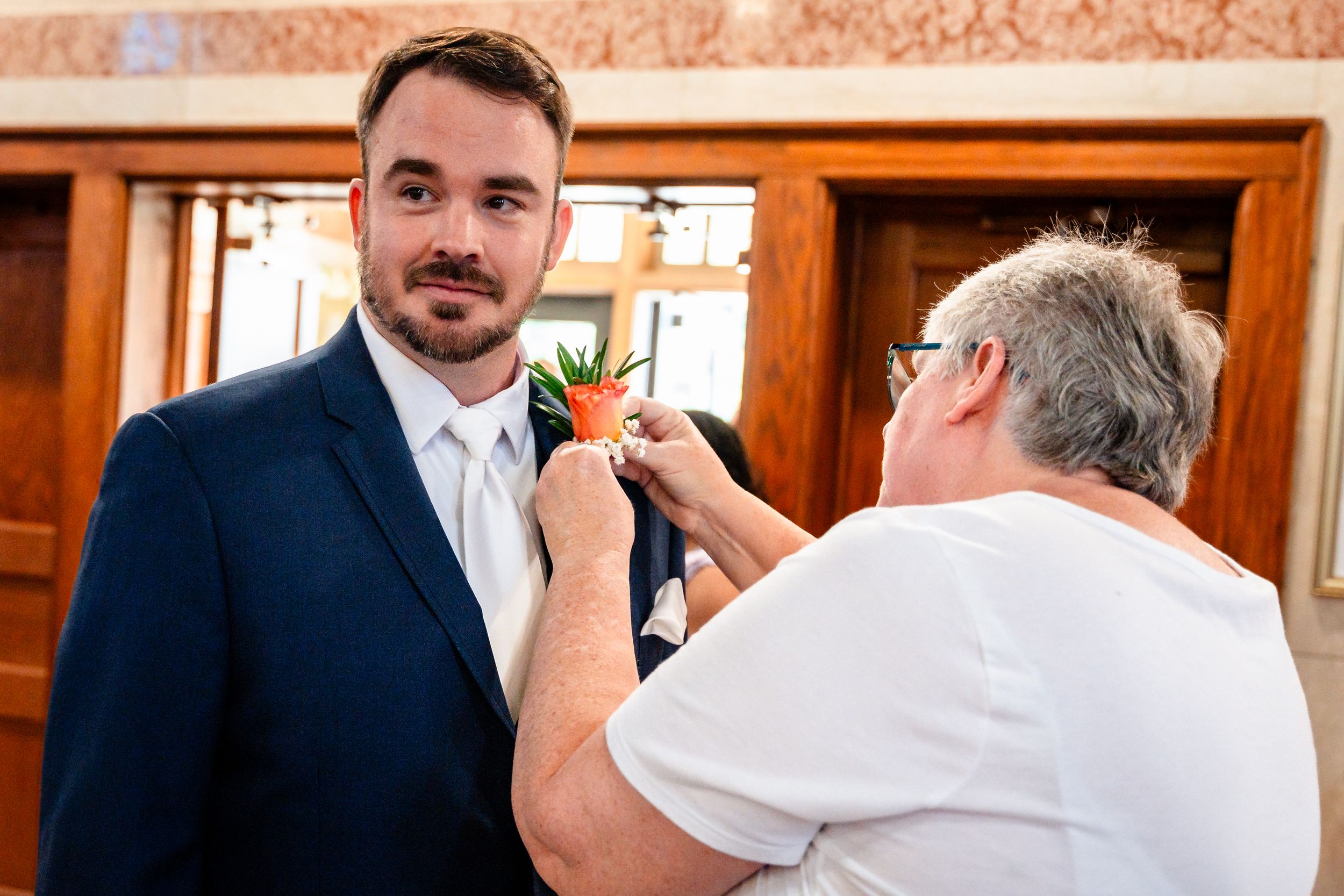Groom in navy suit having boutonniere pinned on lapel by an older woman indoors.