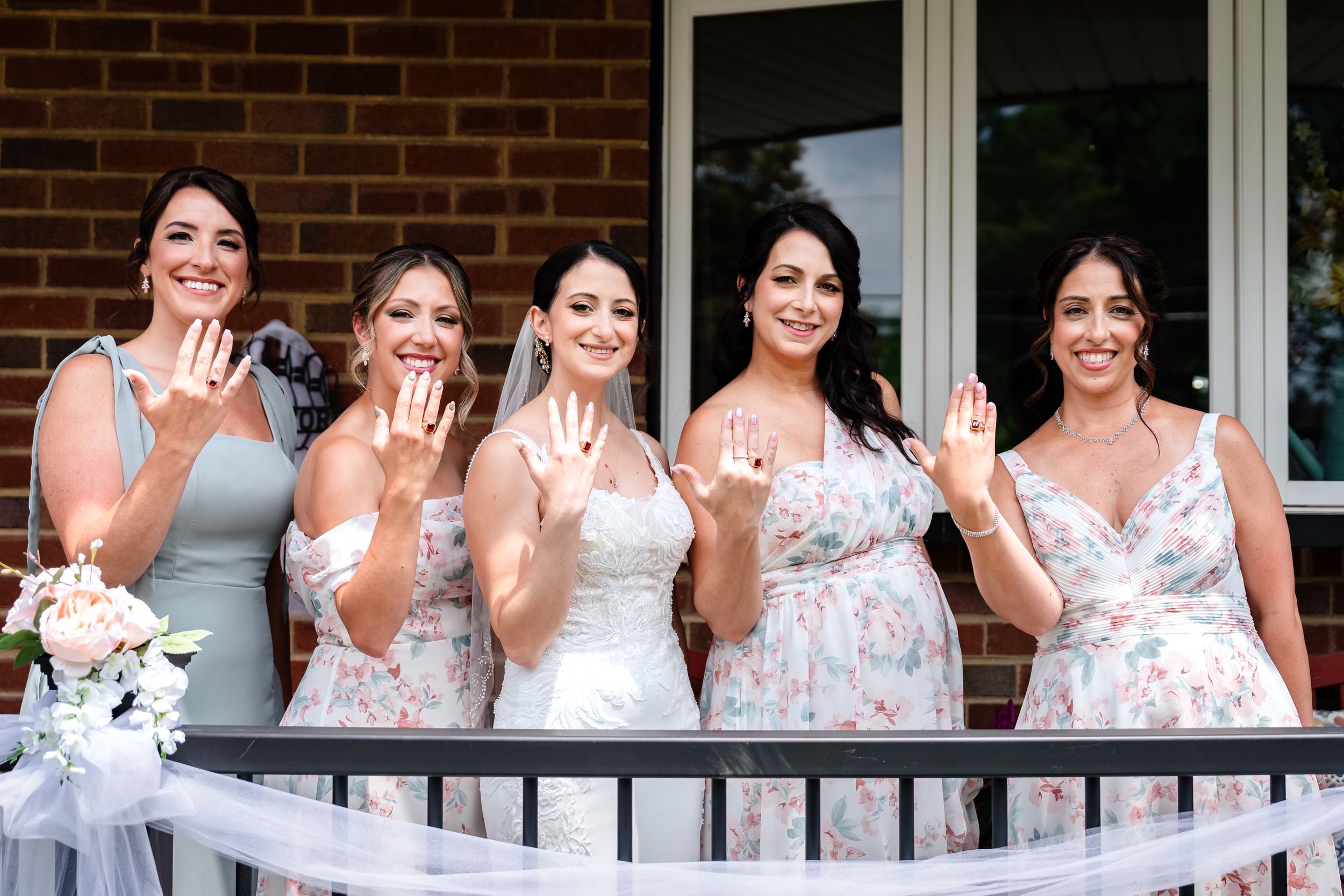 Bride and four bridesmaids in floral dresses show off rings on balcony together.