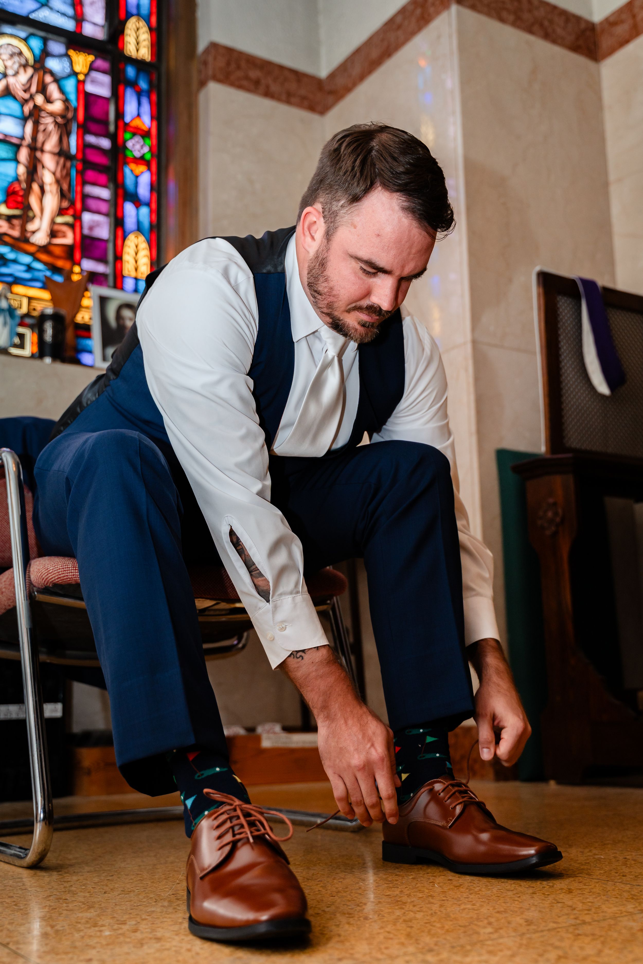 Groom in navy vest and white shirt tying brown dress shoes in a church with stained glass windows.