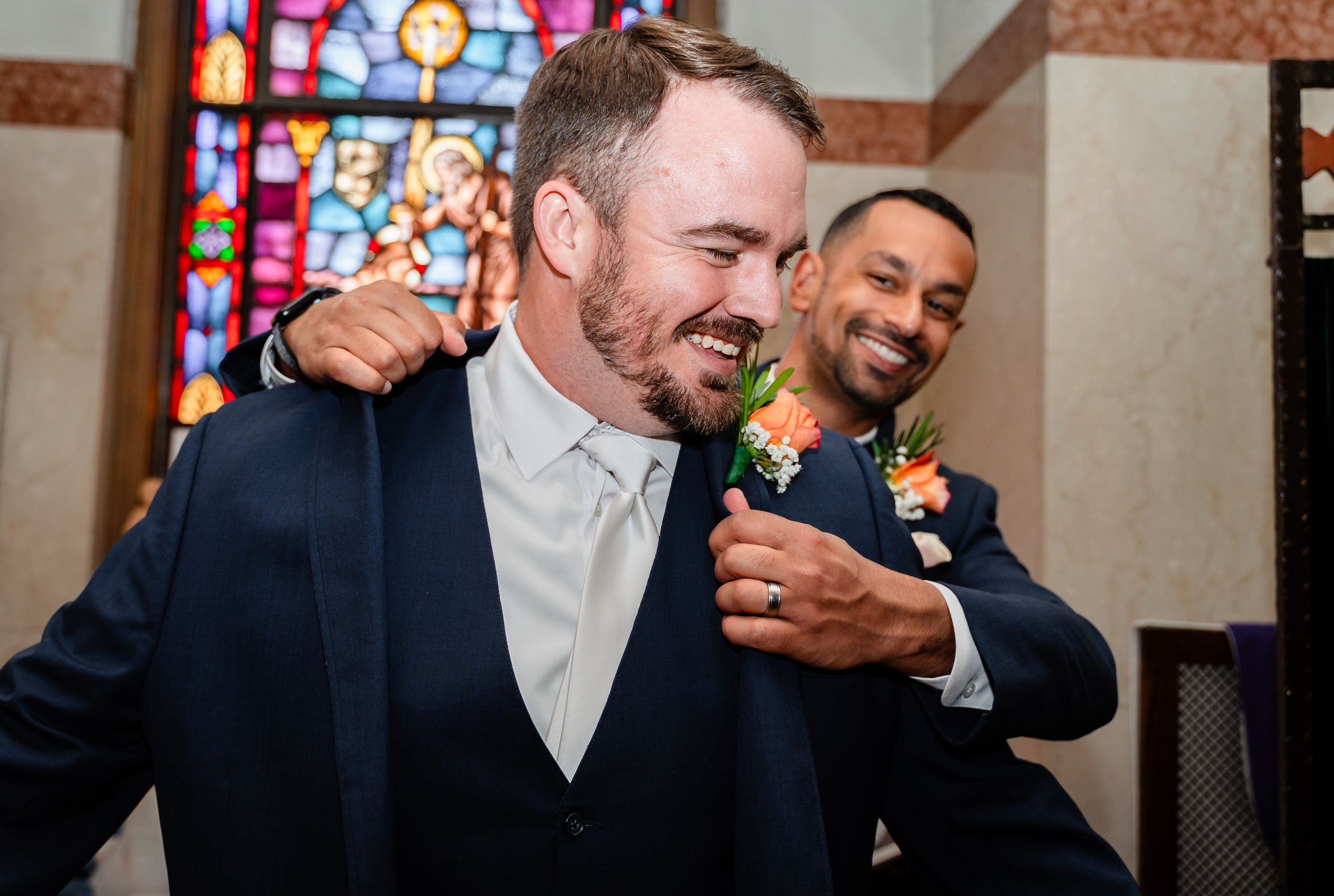 Smiling groom adjusting suit collar with groomsman in church before wedding ceremony.
