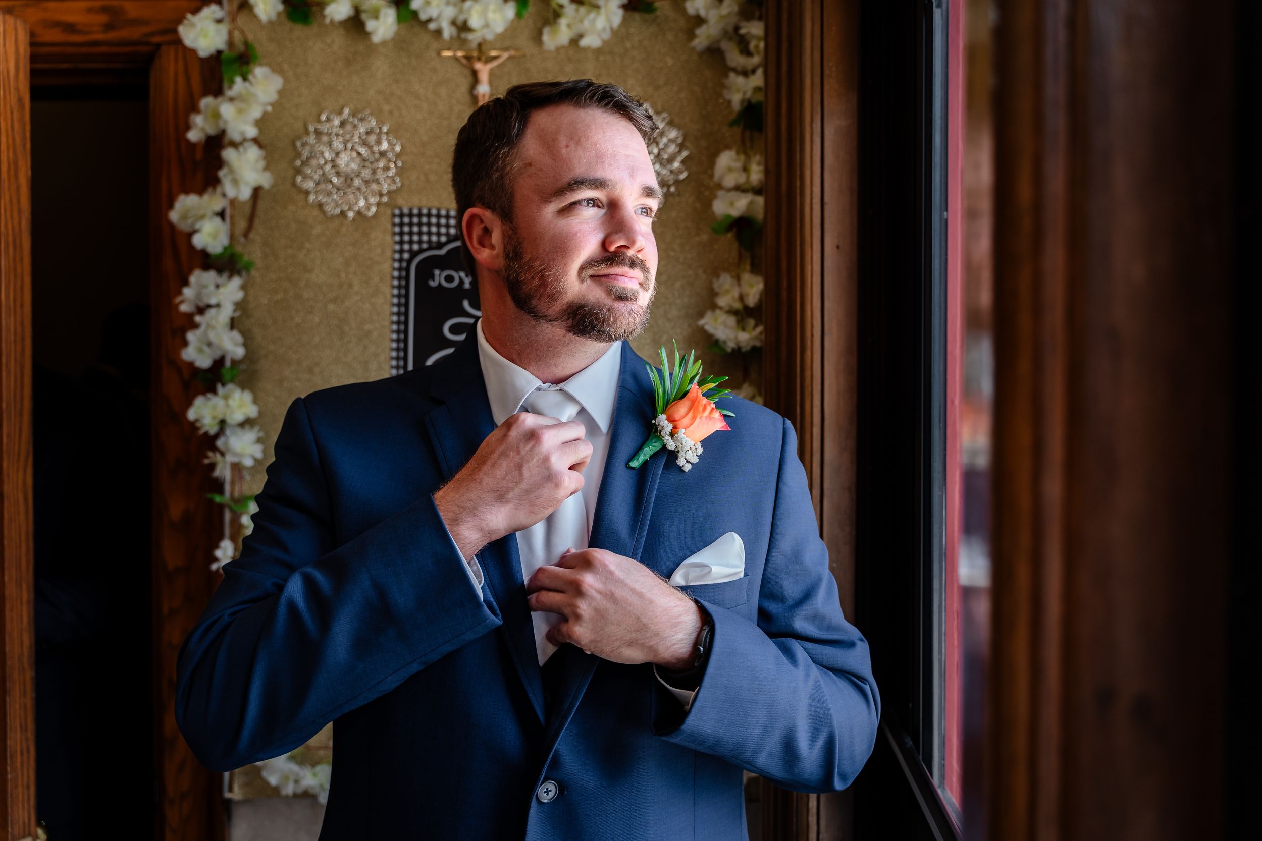 Groom in blue suit fixing his tie near window with floral boutonniere.