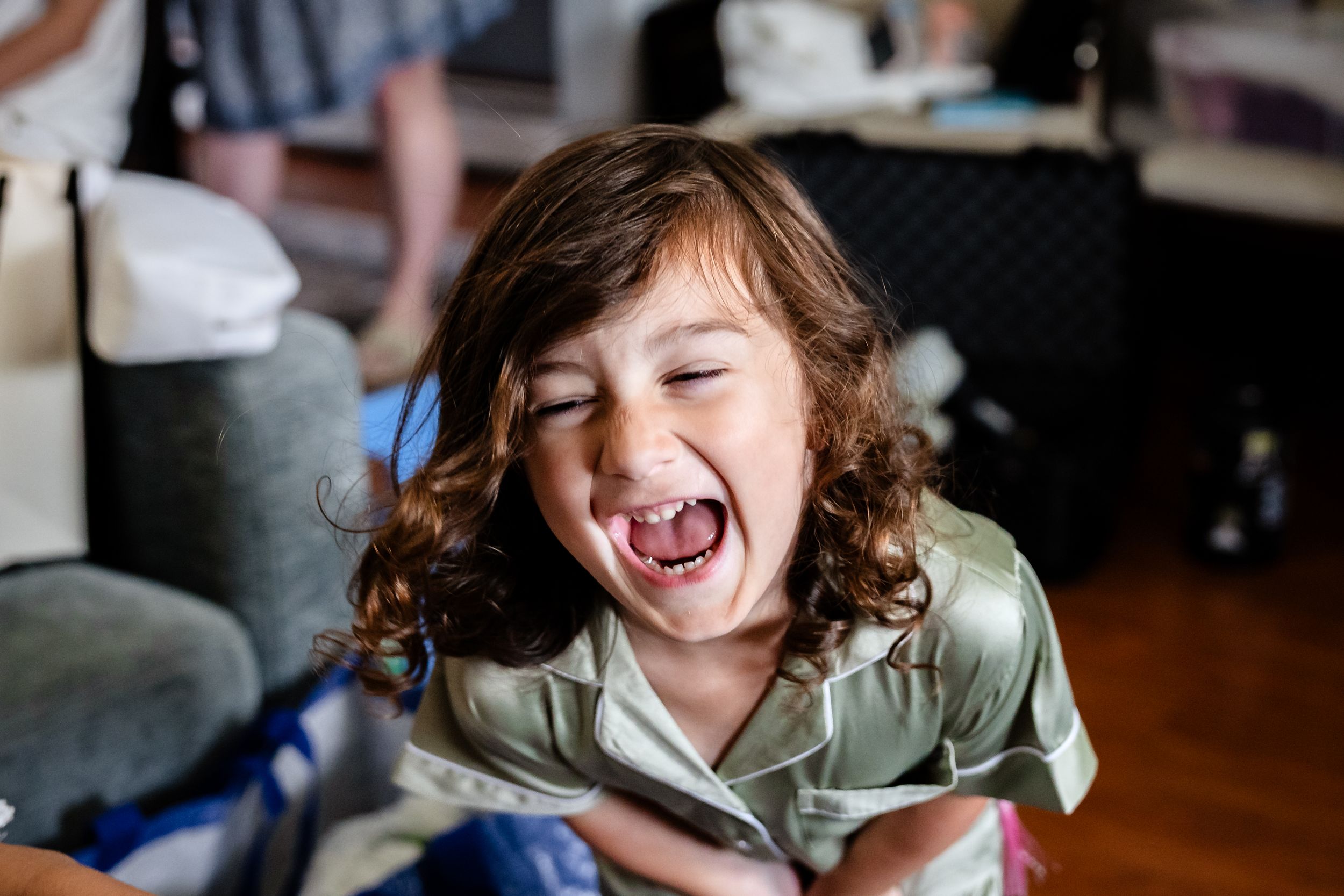 Joyful young child with curly hair laughing with mouth wide open, wearing a satin pajama top indoors.