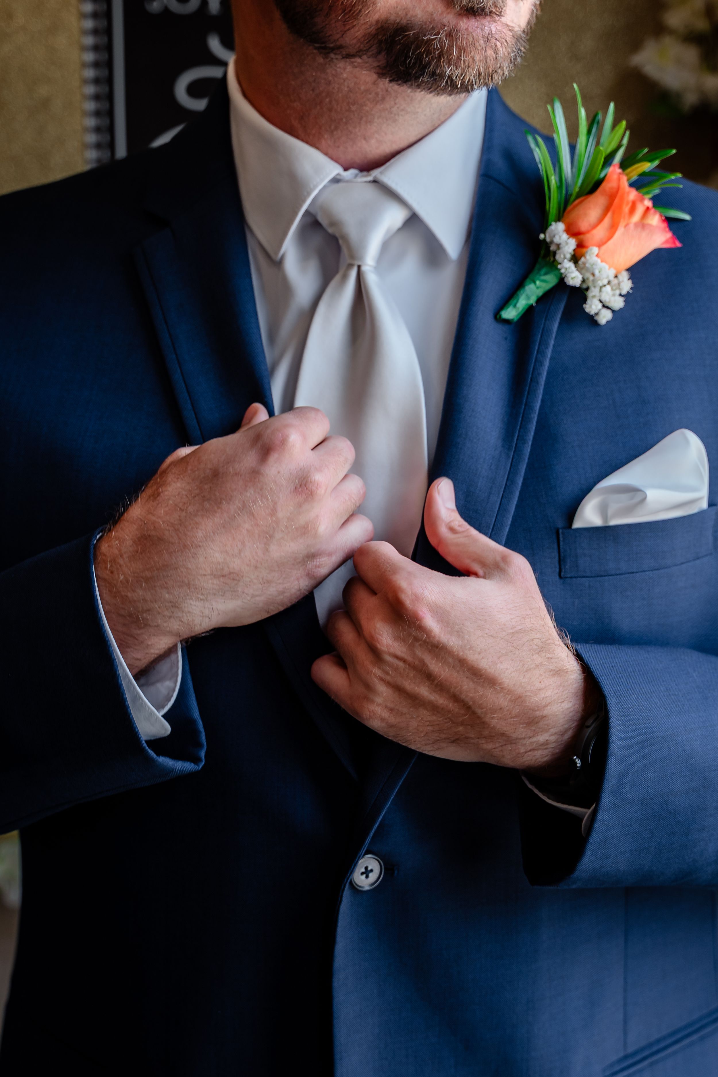 Groom in blue suit adjusting his white tie with orange boutonniere on lapel.