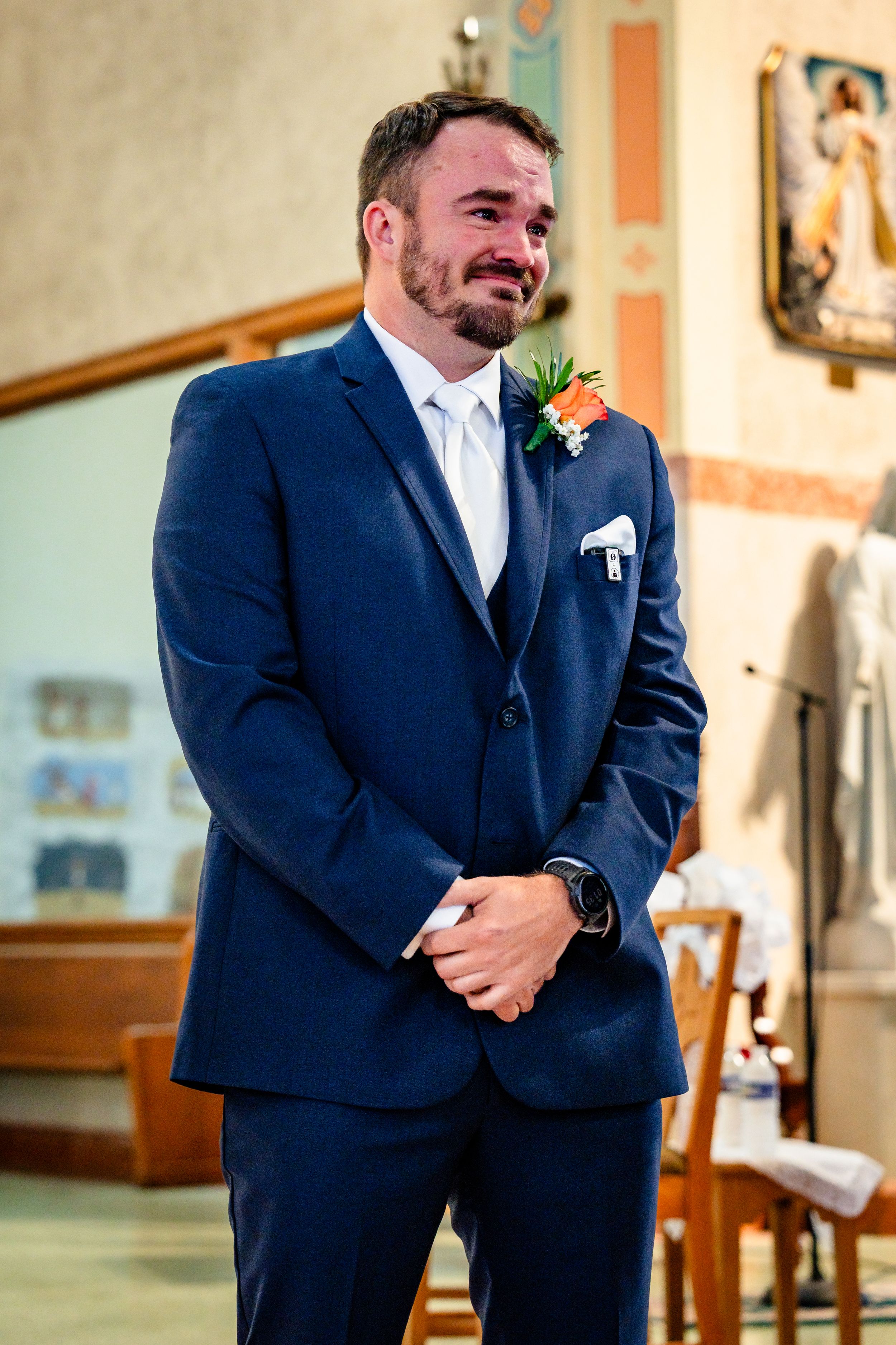 Emotional groom in navy blue suit with boutonniere crying at the altar during wedding ceremony in church.