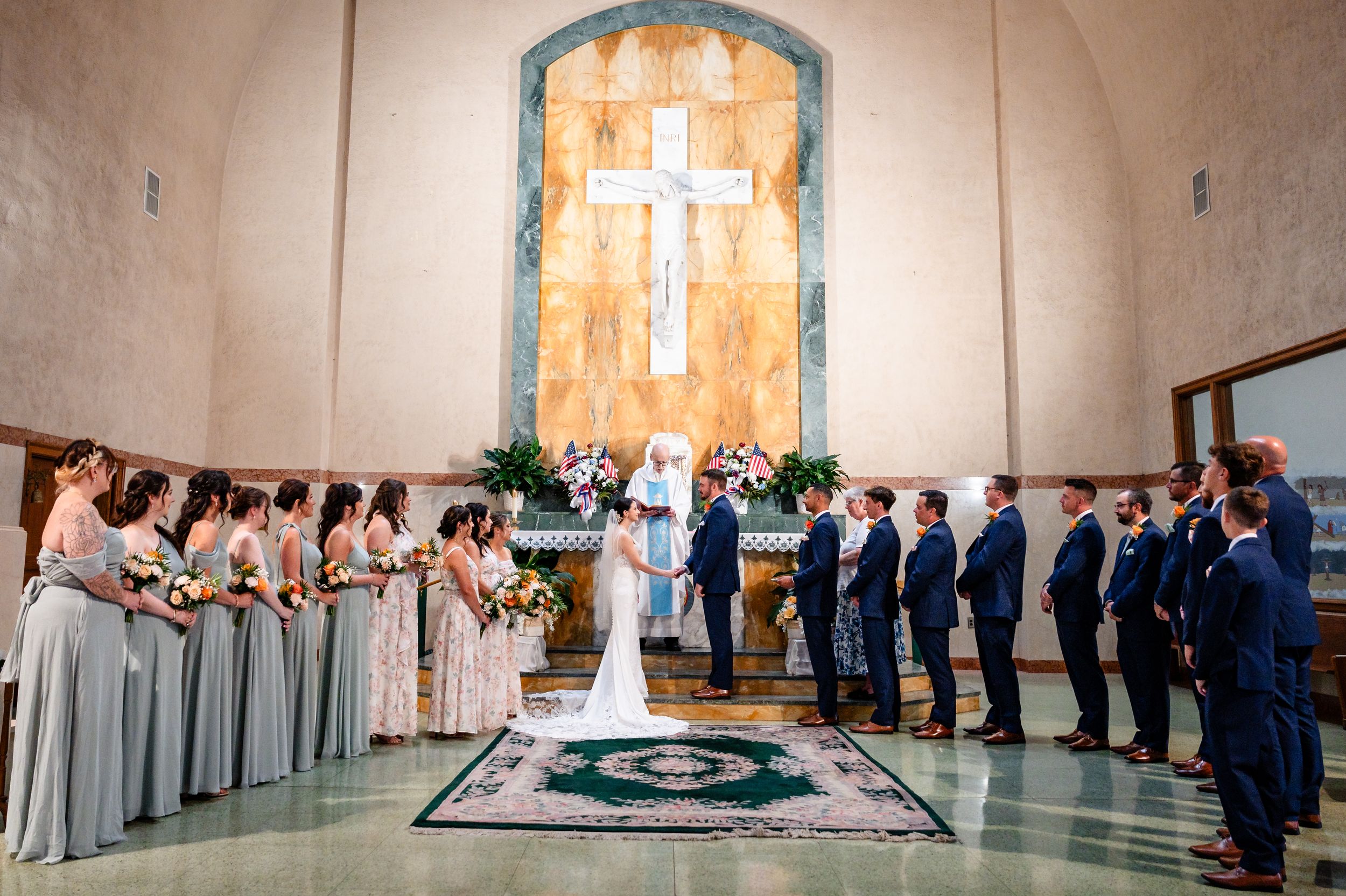 Wedding ceremony in a church with bridesmaids in sage dresses and groomsmen in navy suits before an altar.