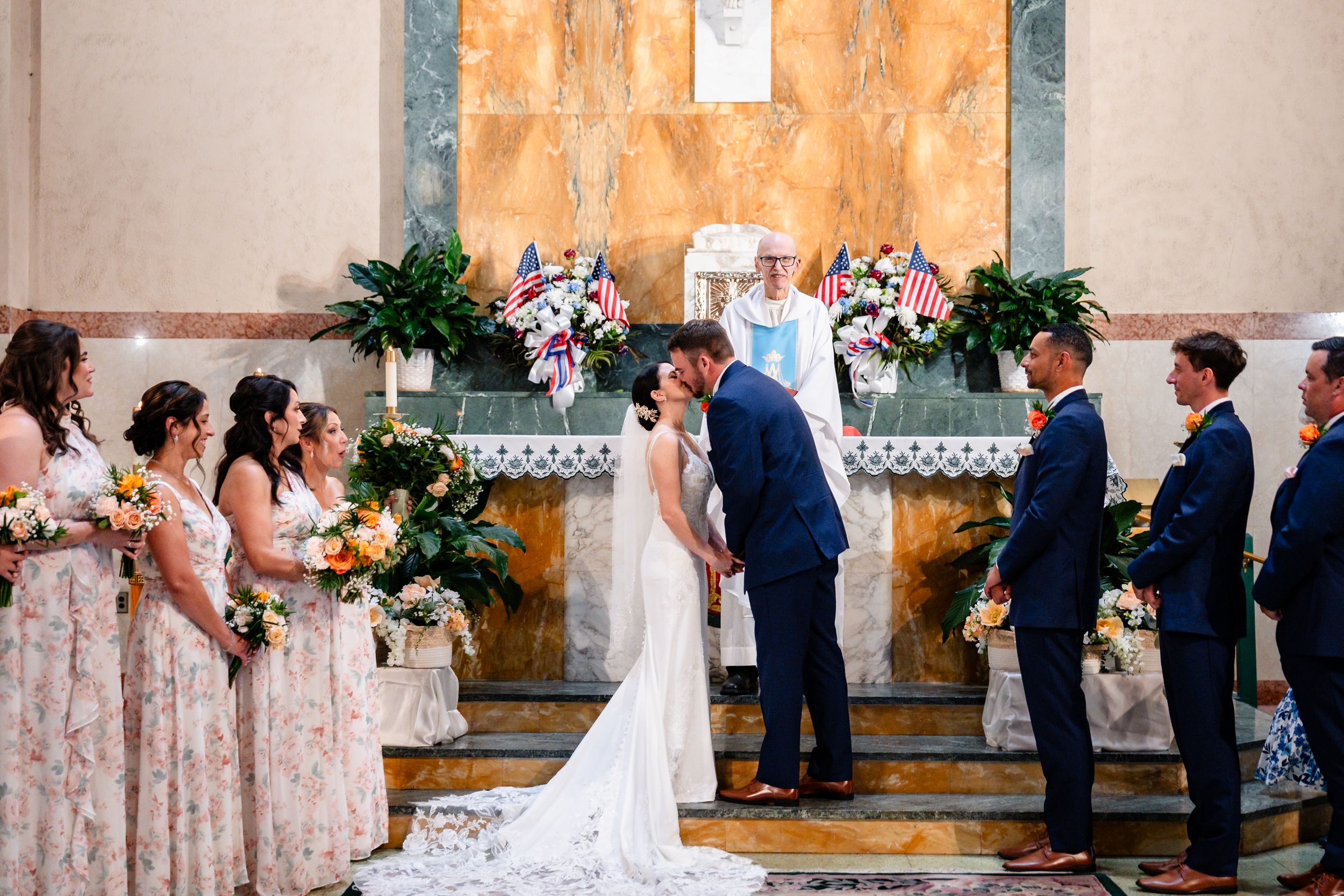 Bride and groom sharing first kiss at altar surrounded by wedding party.
