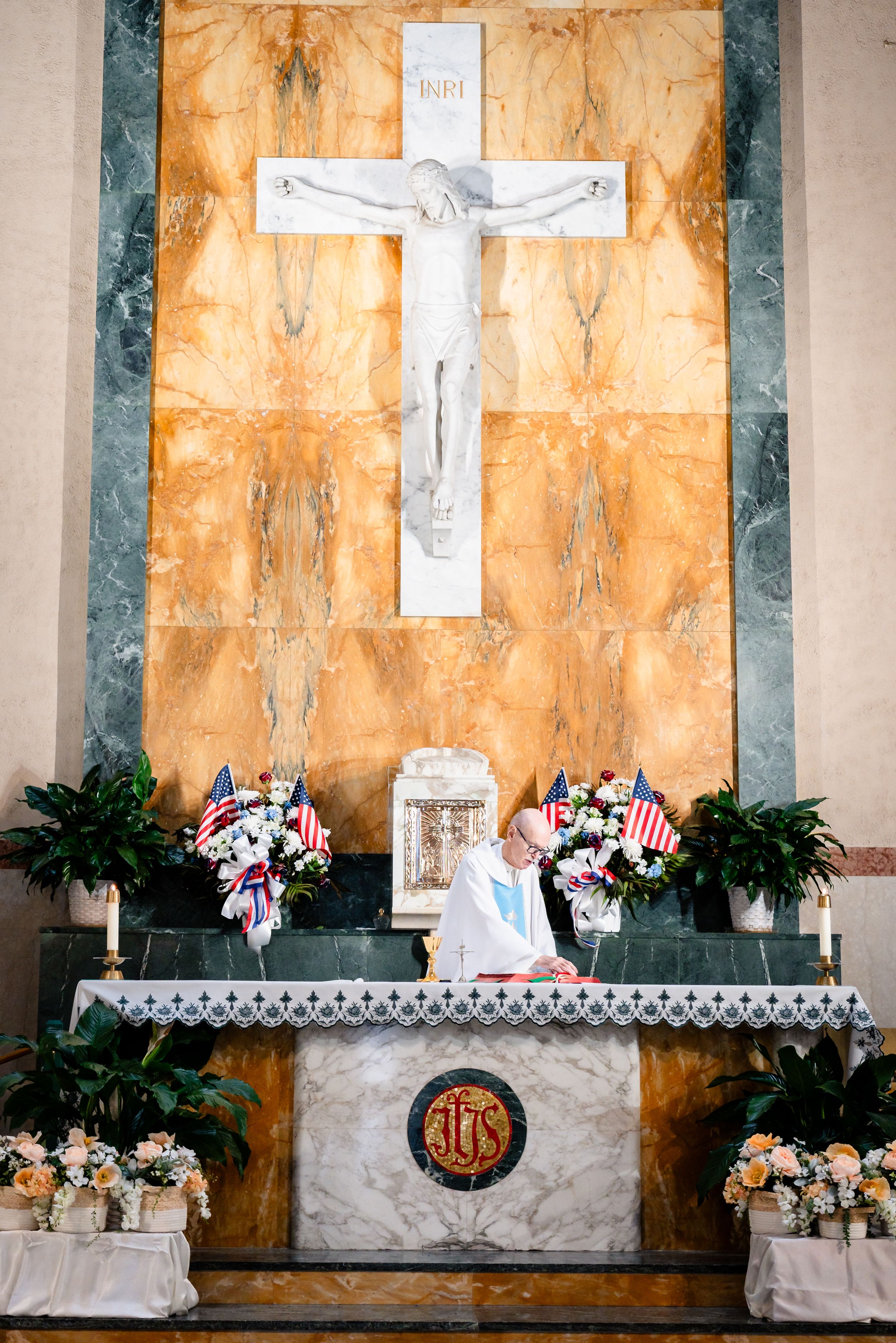 Priest kneeling at ornate church altar with large marble crucifix, American flags, and floral arrangements during a ceremony.