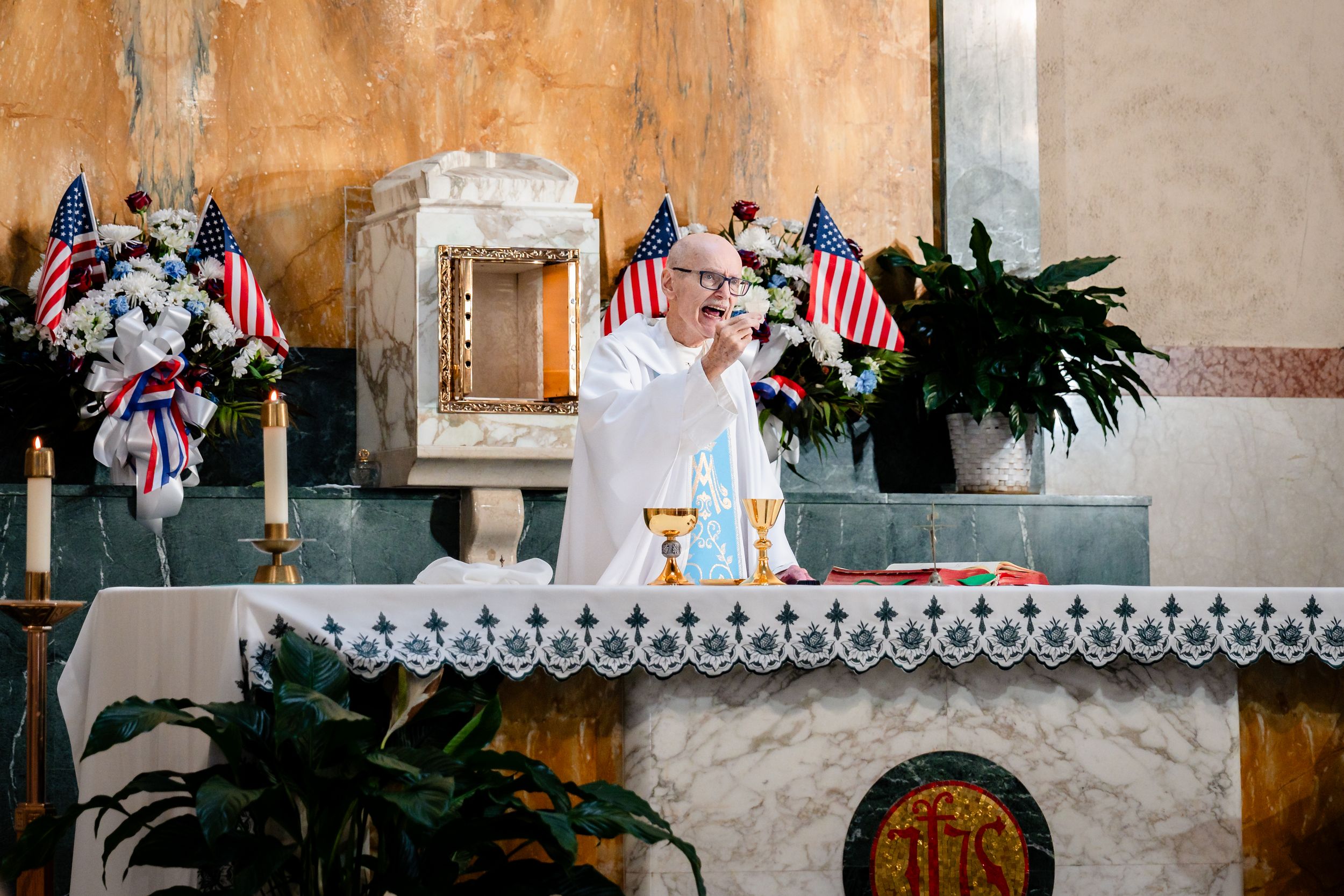Priest performs wedding ceremony at decorated altar with American flags and floral arrangements.