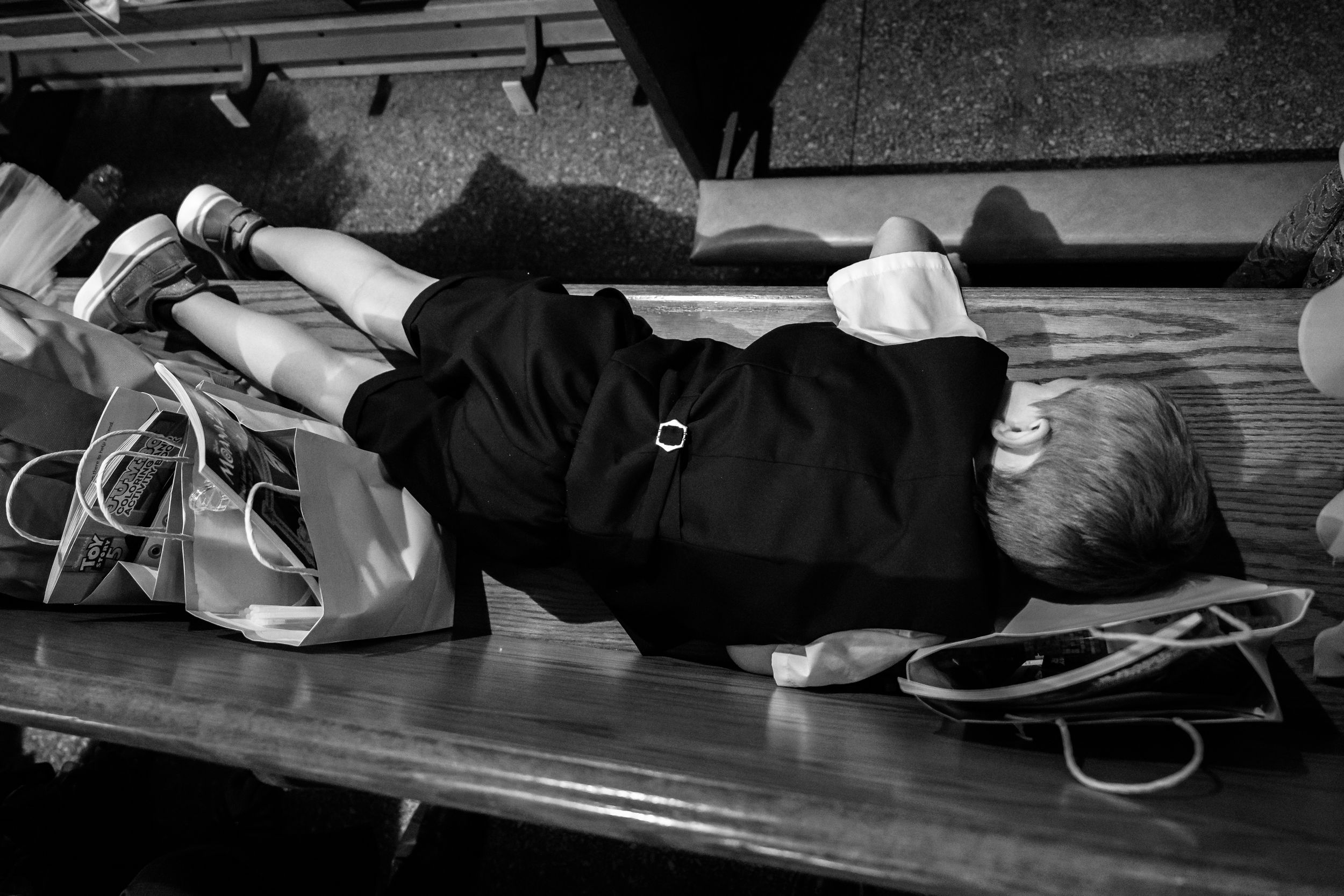 Black and white photo of a person lying on a wooden bench with bags beside them.