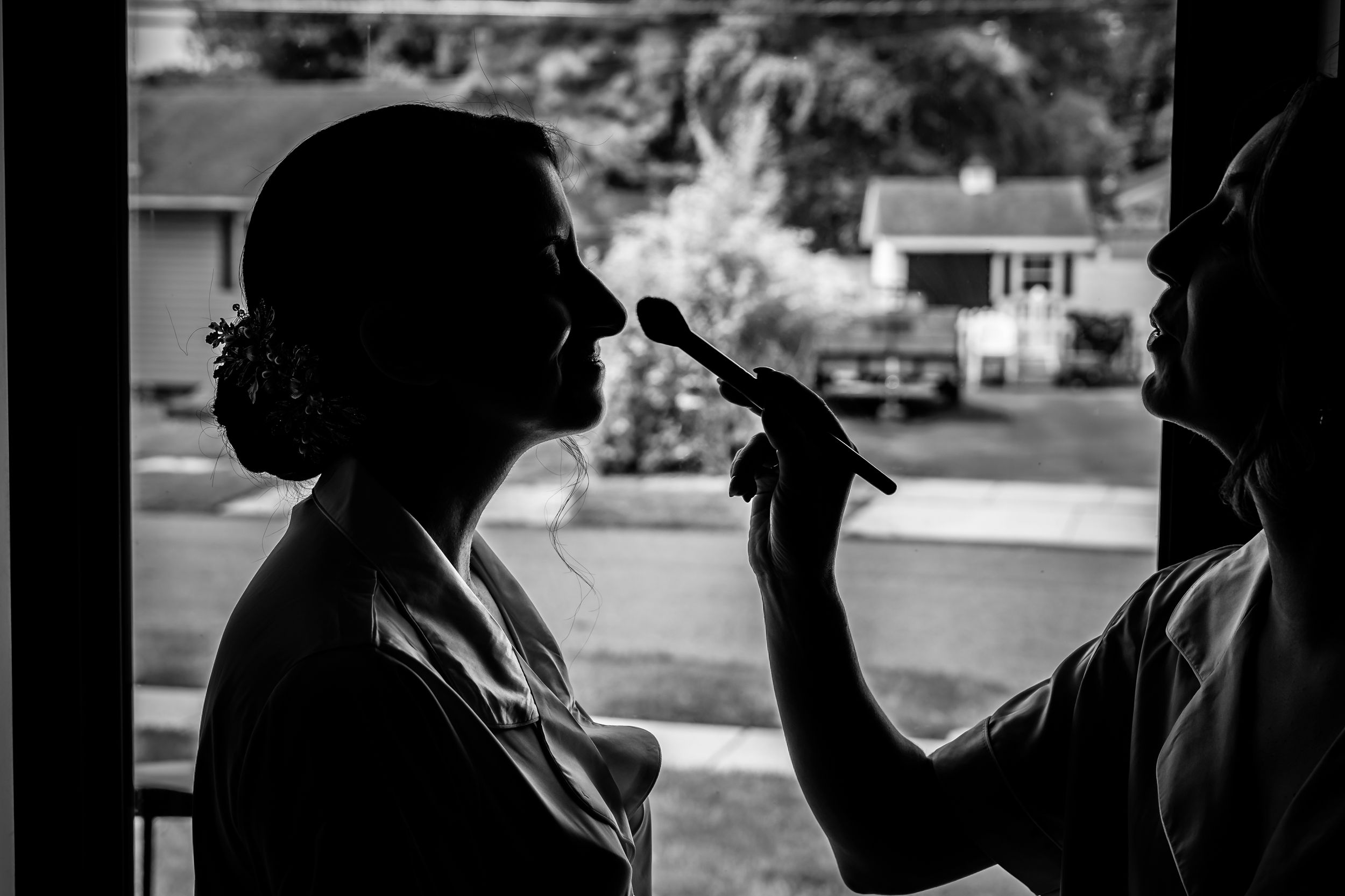 Black and white silhouette of a makeup artist applying makeup to a bride near a window before her wedding.